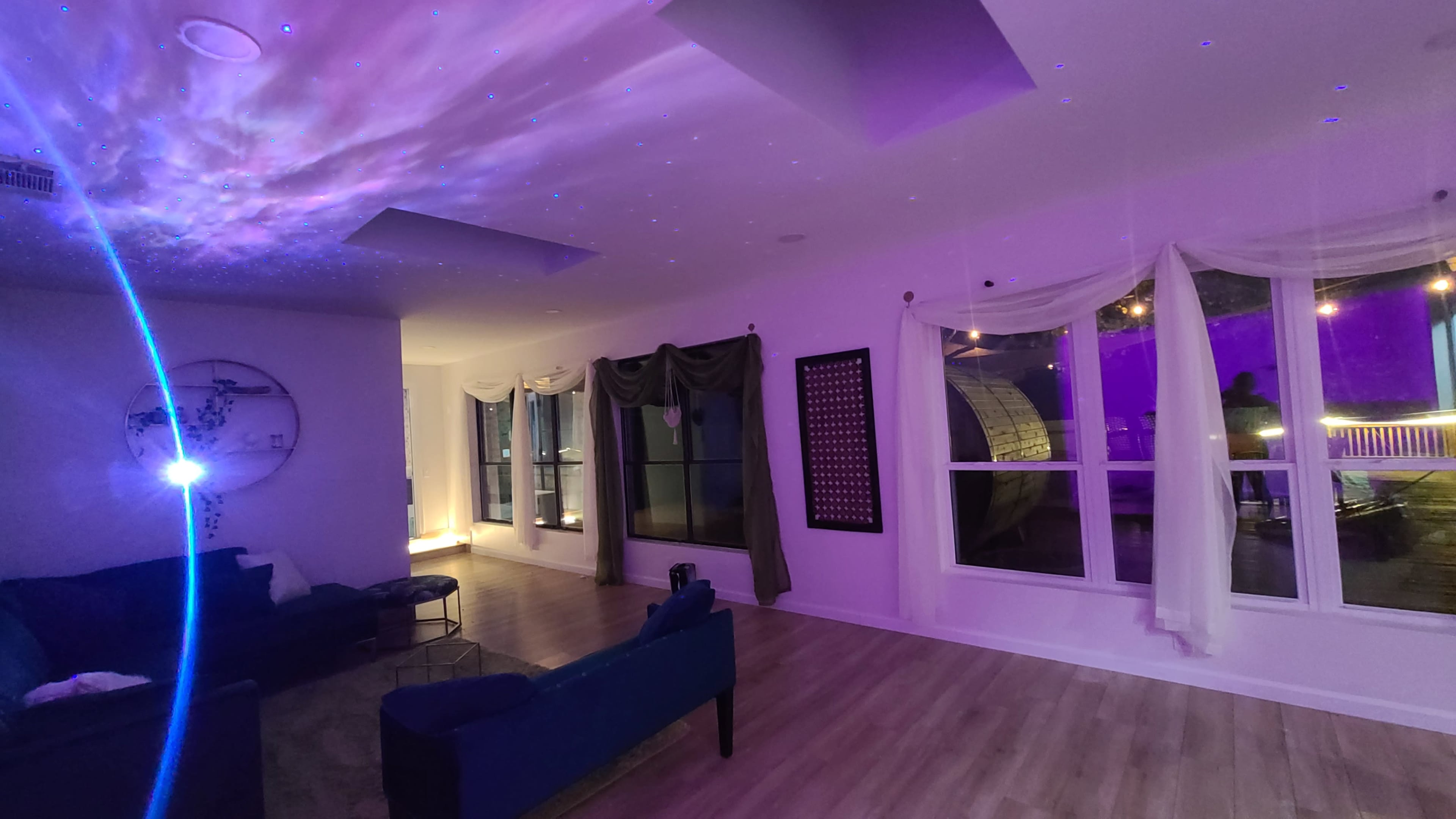 The image shows a spacious living room with a dark blue couch, large windows illuminated by purple light, and a ceiling displaying a starry projection.
