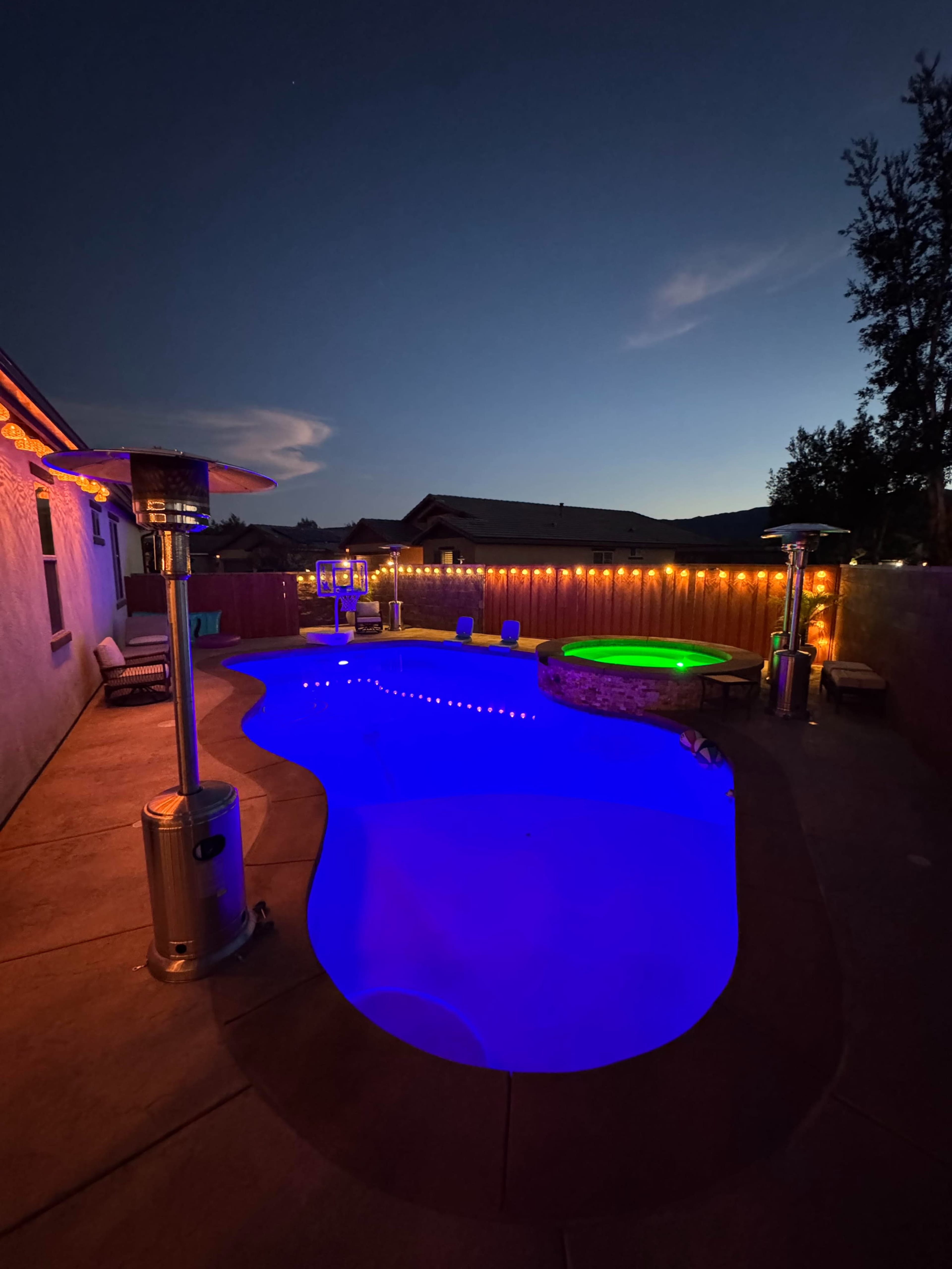 A brightly lit swimming pool with colorful LED lights and patio heaters is set against a twilight sky.