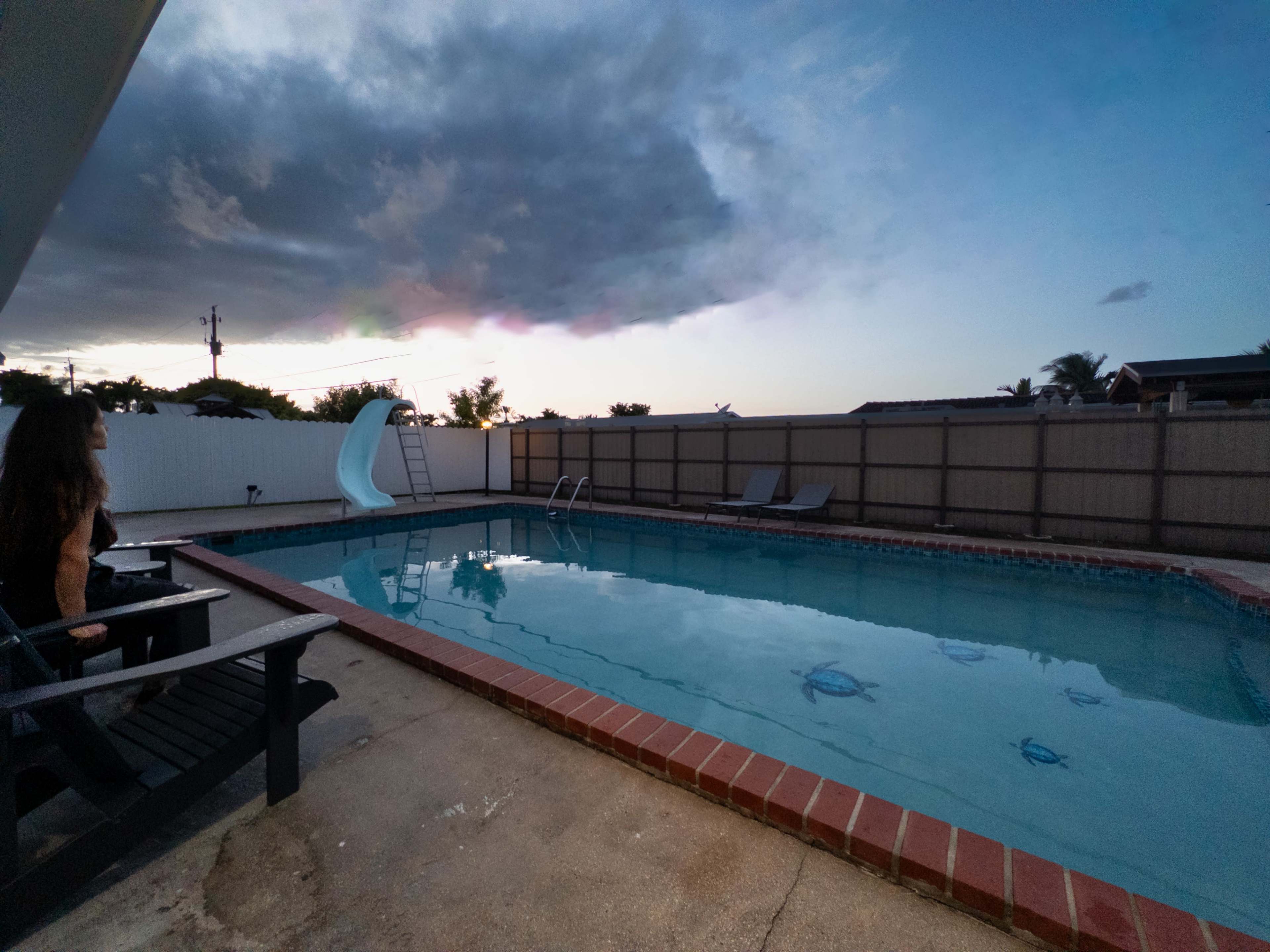 A swimming pool with a slide is surrounded by a concrete deck, and a person is sitting on a chair, facing the water as clouds gather in the sky.