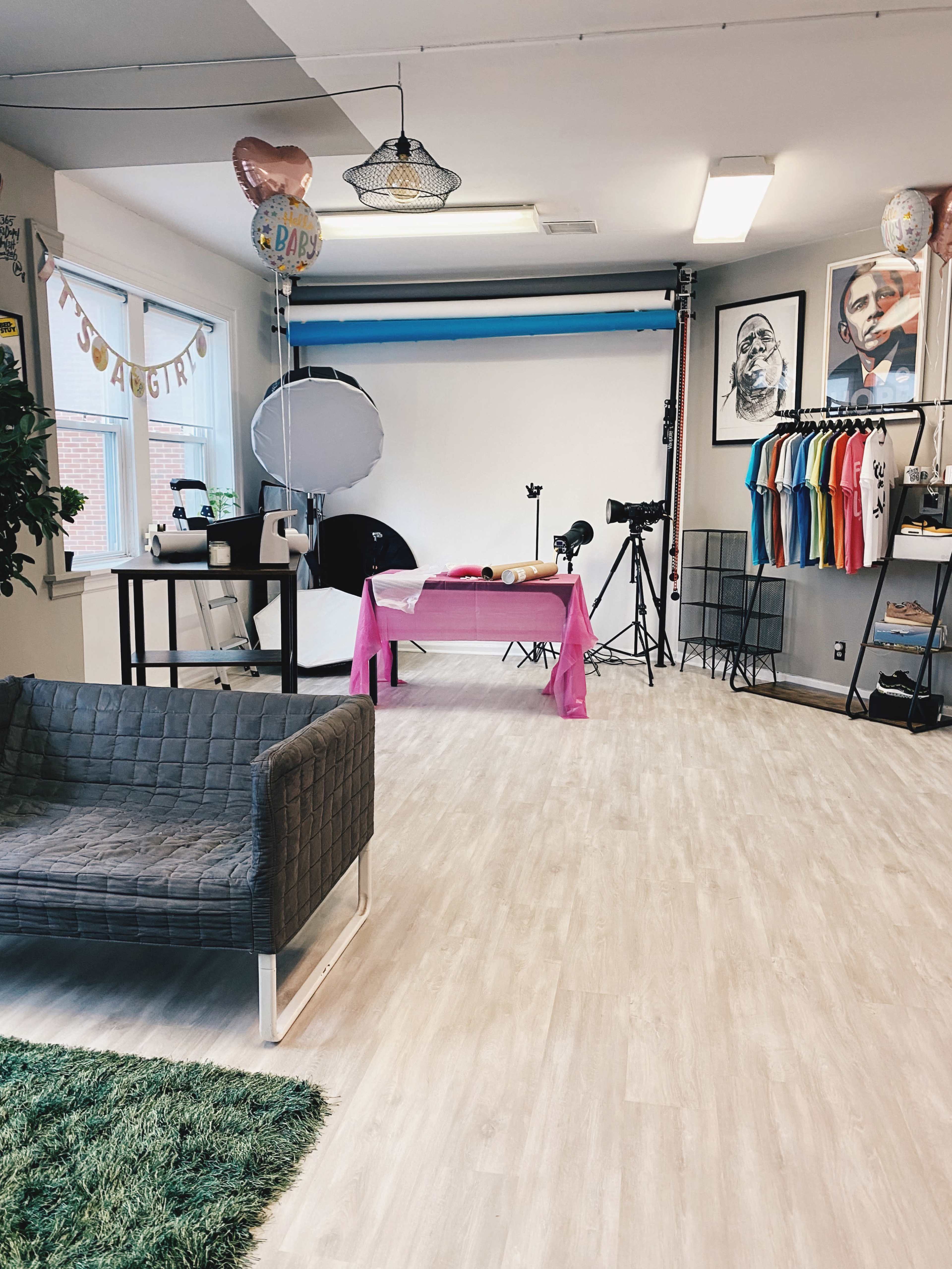 A studio space with a pink tablecloth, photography equipment, a gray couch, and colorful shirts hanging on one wall.