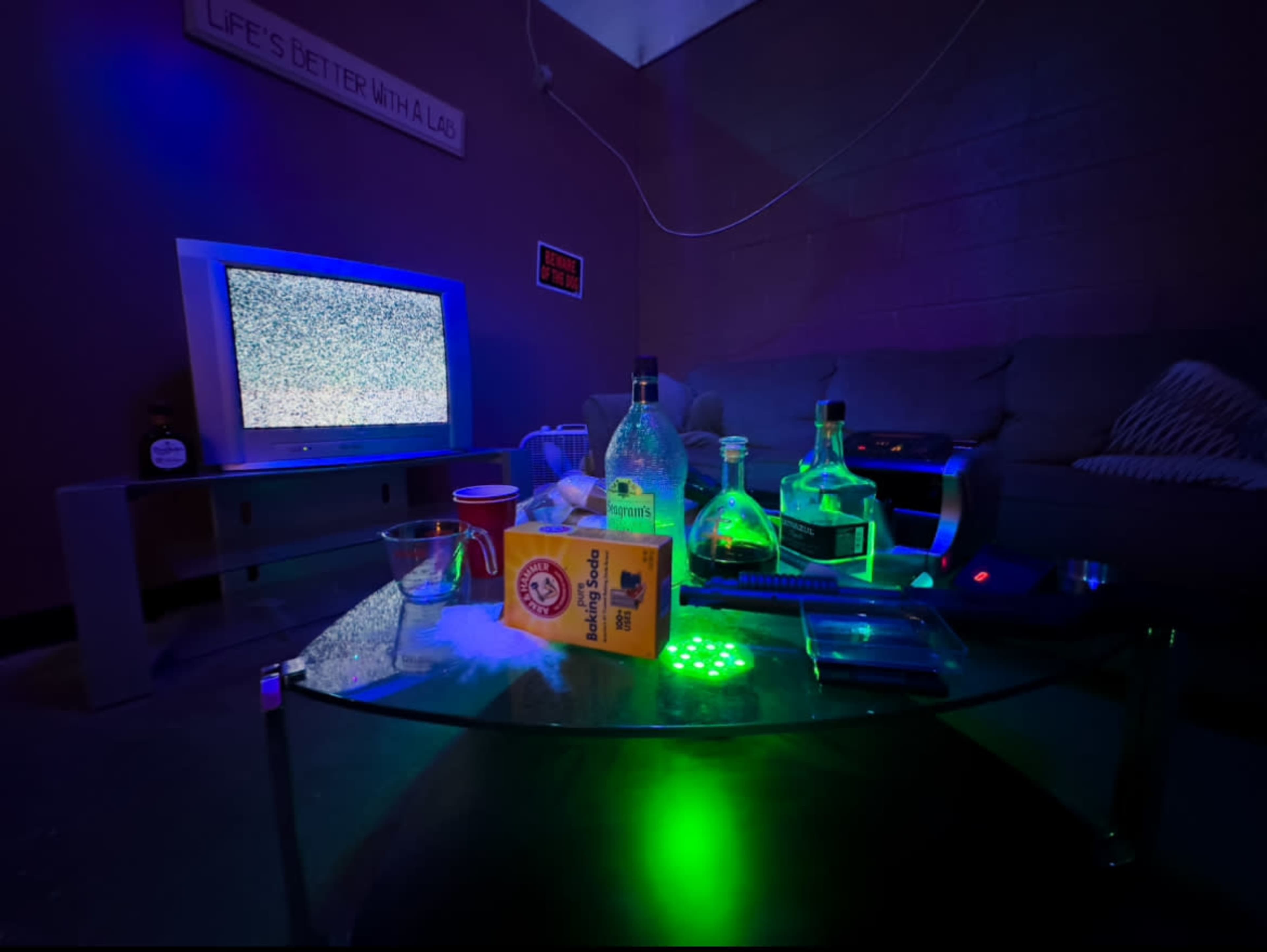 A living room with a vintage TV displaying static, a glass coffee table littered with bottles, a cup, and a box, and a couch in the background illuminated by green lighting.
