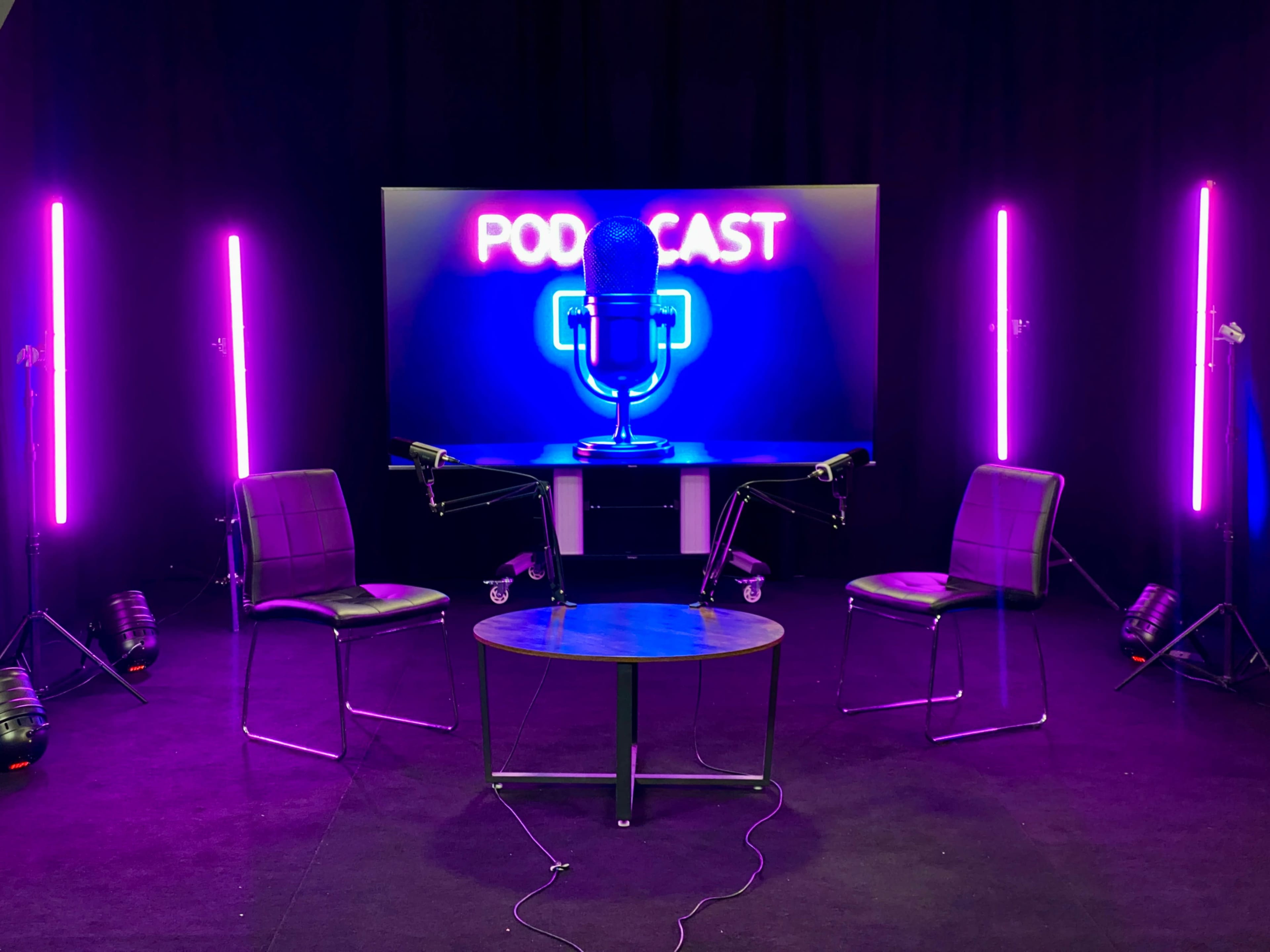 The image features a podcast studio setup with two chairs, a round table, and a colorful neon backdrop displaying the word "PODCAST."