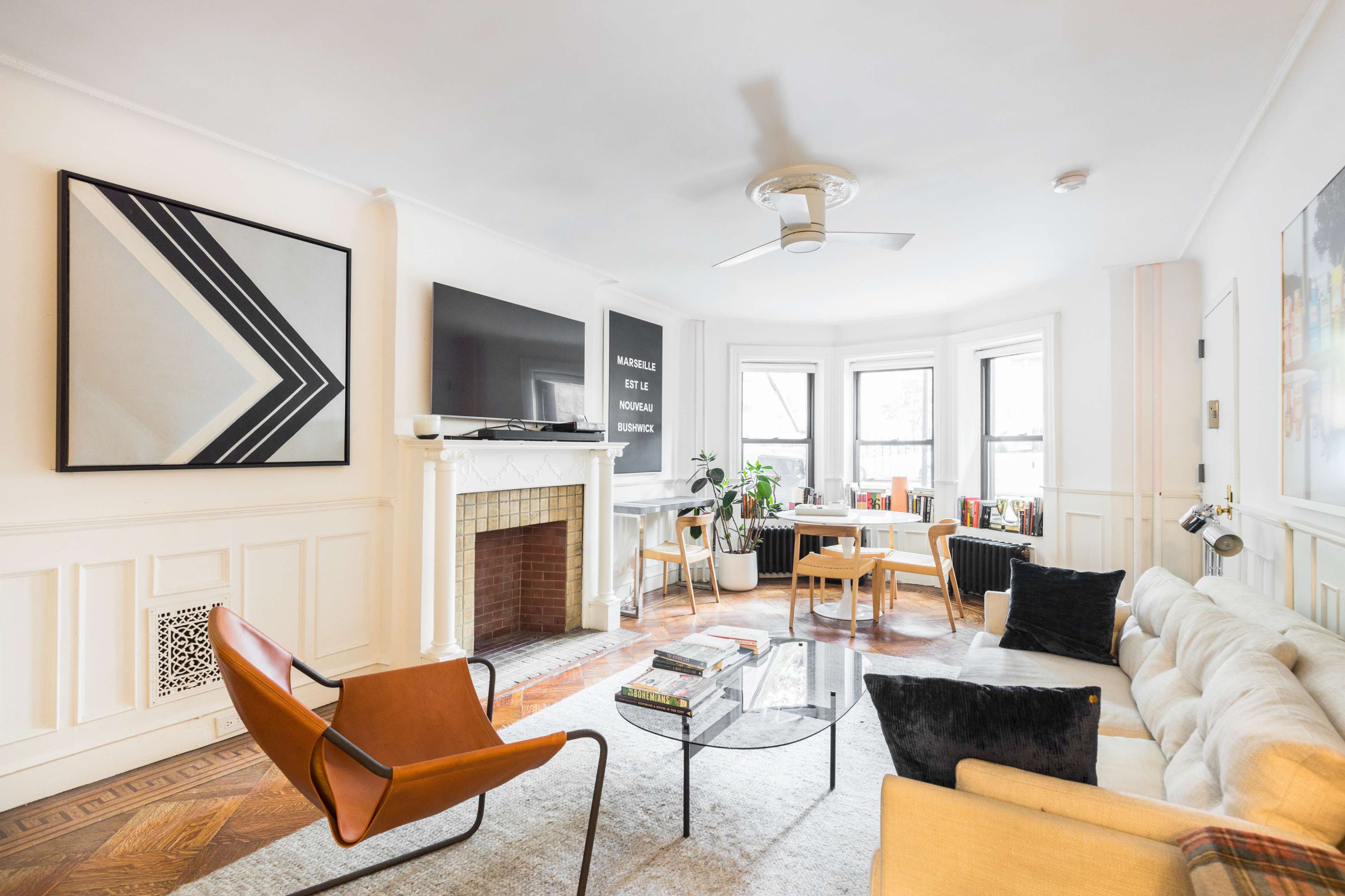A modern living room features a beige sofa, an orange chair, a glass coffee table, and large windows with natural light illuminating the space.