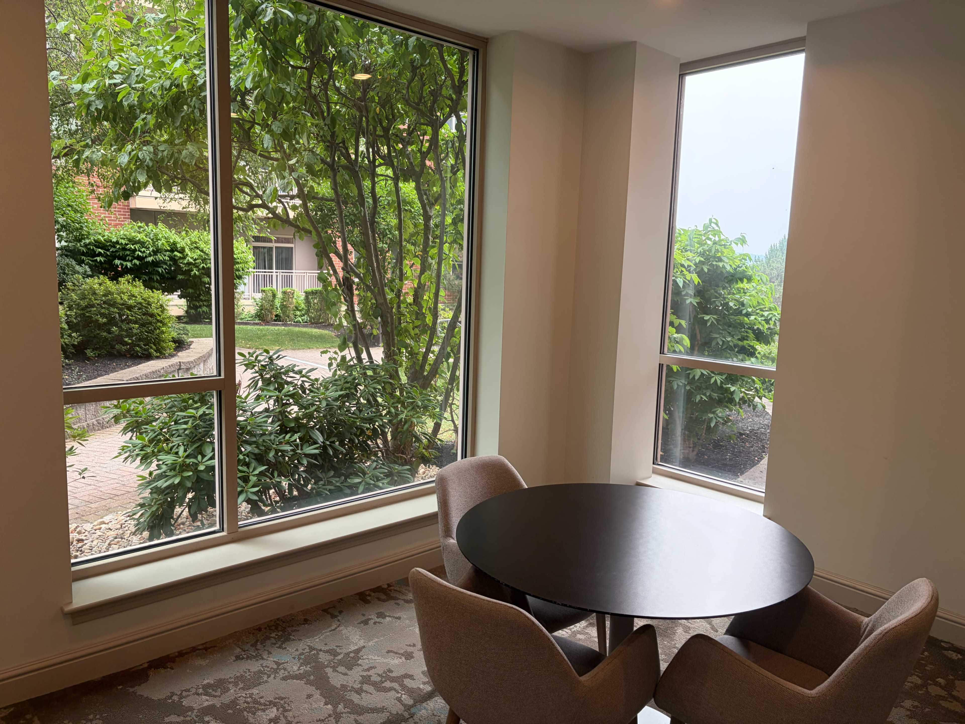 A round table with two chairs is positioned near large windows overlooking a landscaped outdoor area with greenery.