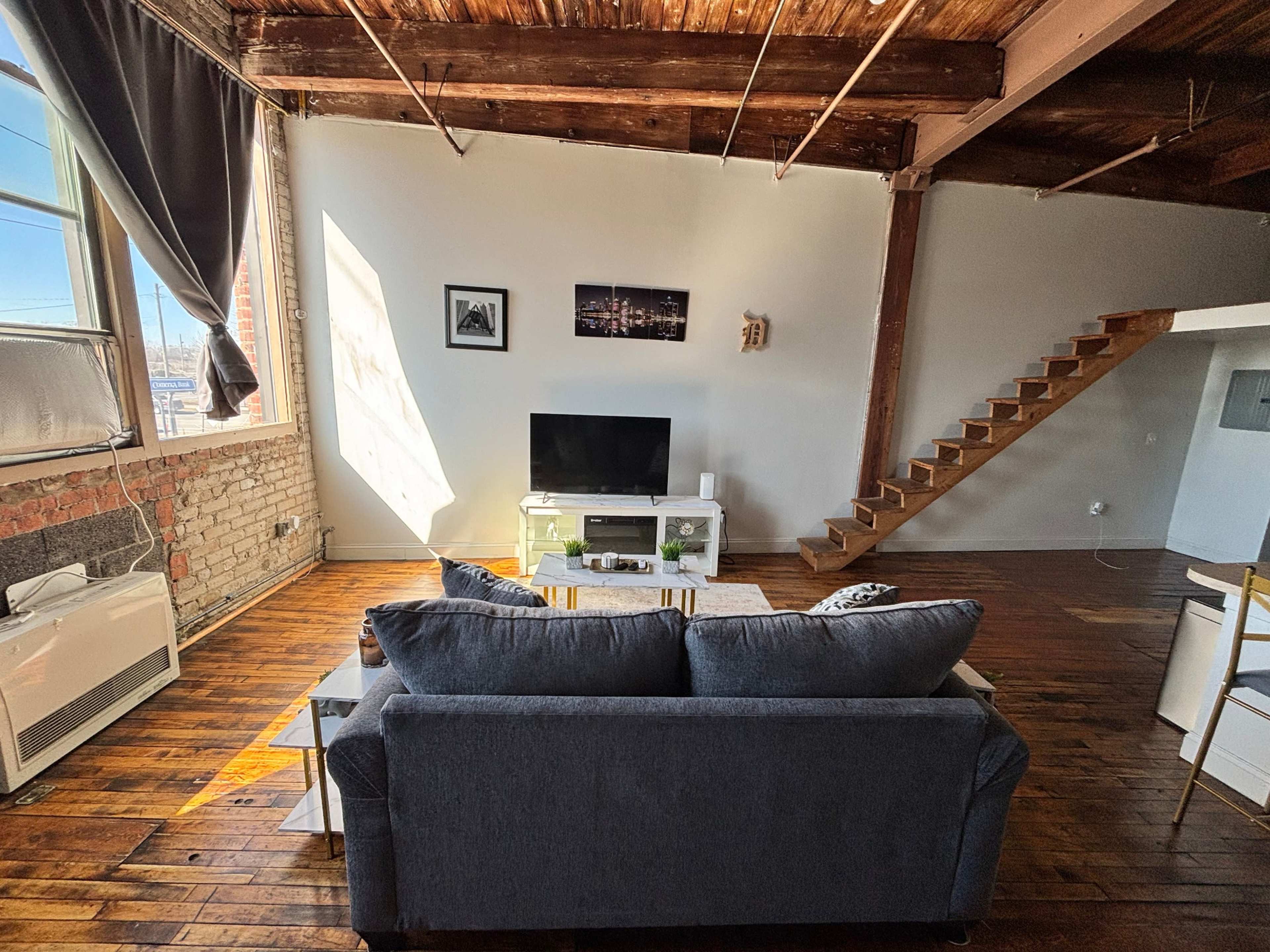 The image shows a modern living room with a gray sofa, a television on a white cabinet, and wooden floors, along with a staircase leading to an upper level.