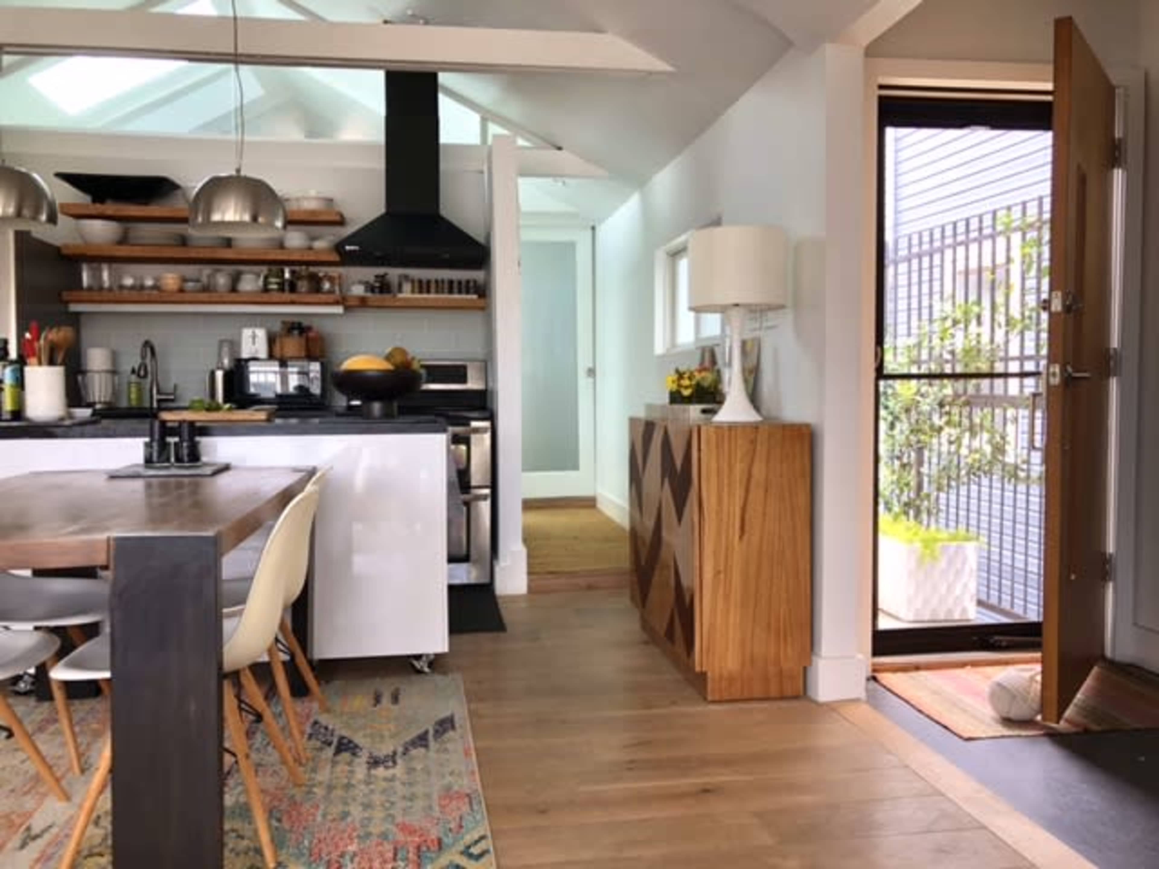 The image shows a modern kitchen and dining area with a wooden dining table, open shelving, and a front door leading to an exterior space.
