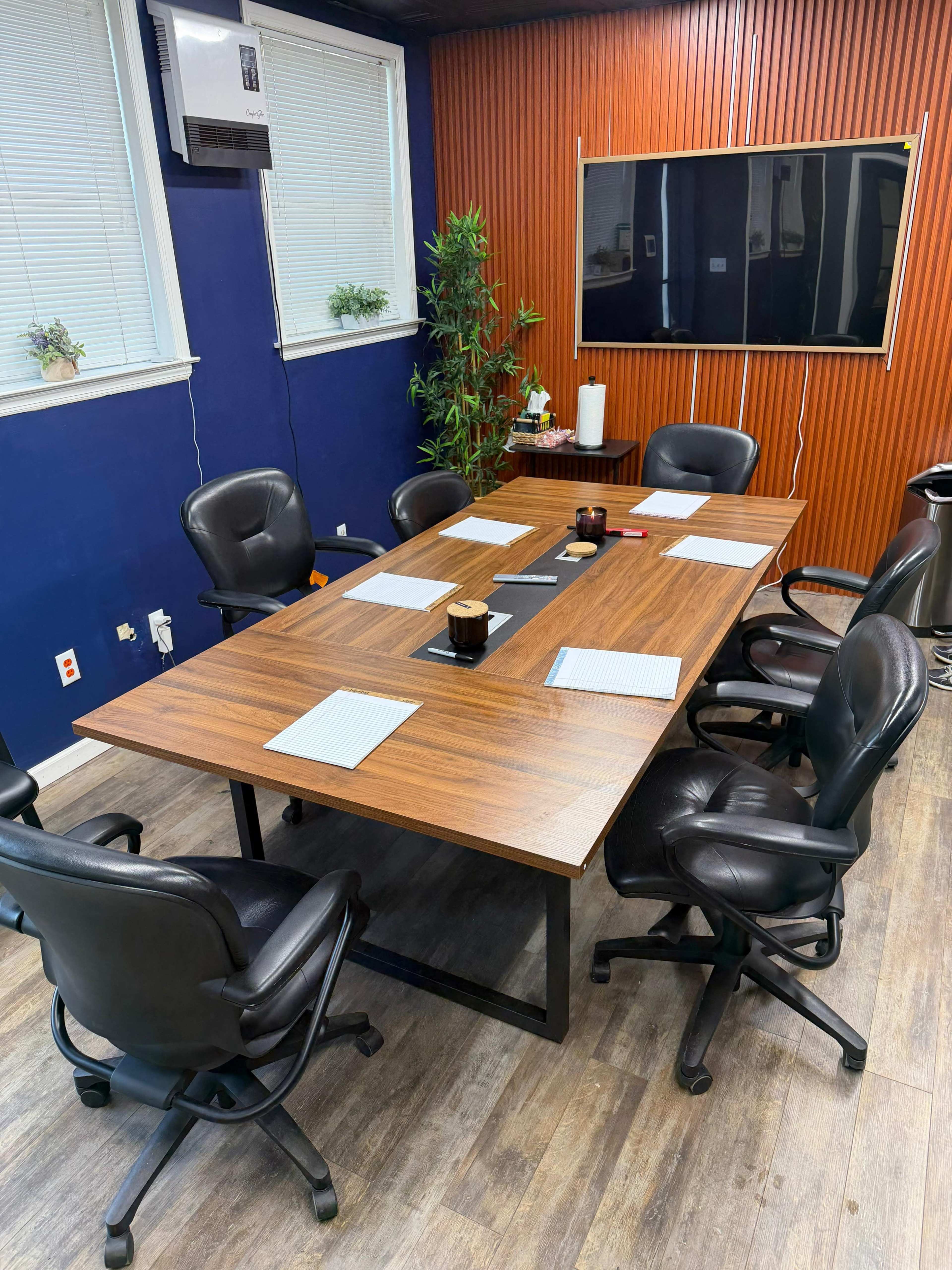 The image shows a conference room with a rectangular wooden table surrounded by black office chairs, a wall-mounted screen, and plants in the background.