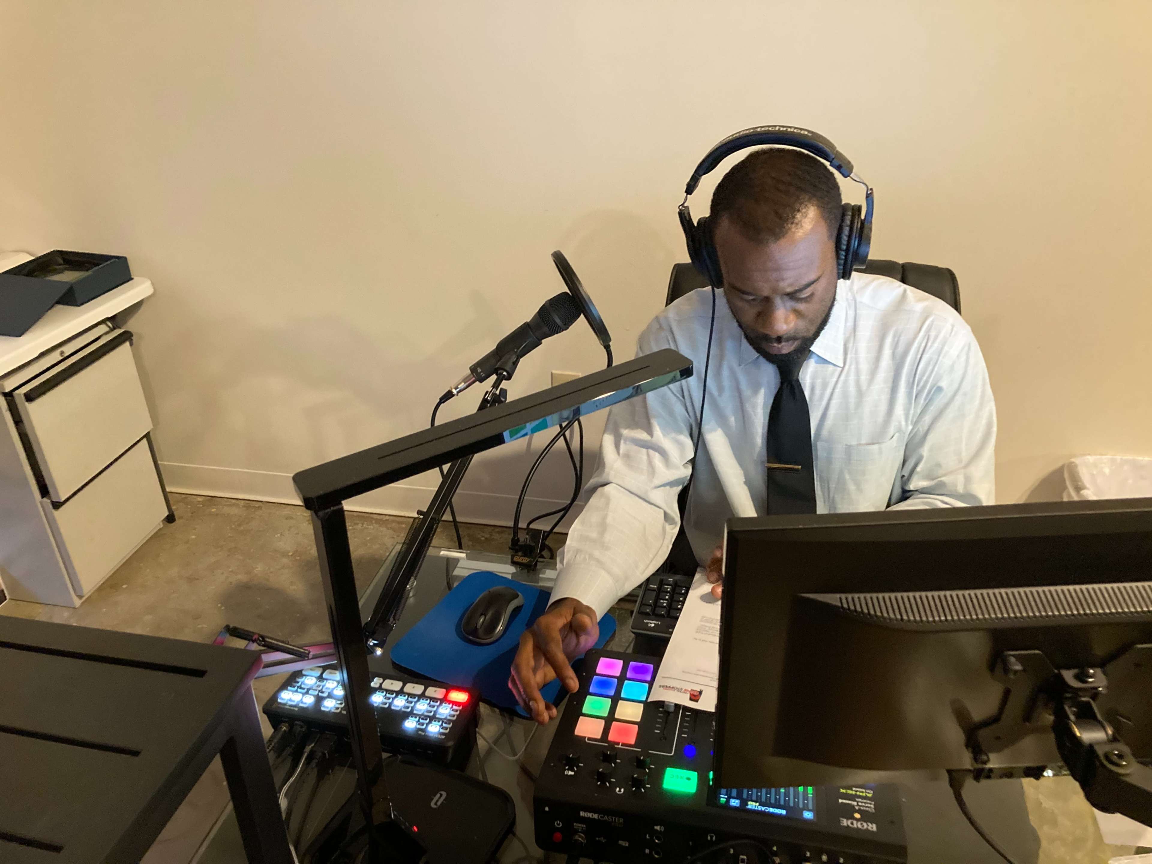 A man in a dress shirt and tie operates a control panel with various buttons and monitors while wearing headphones in a recording studio.