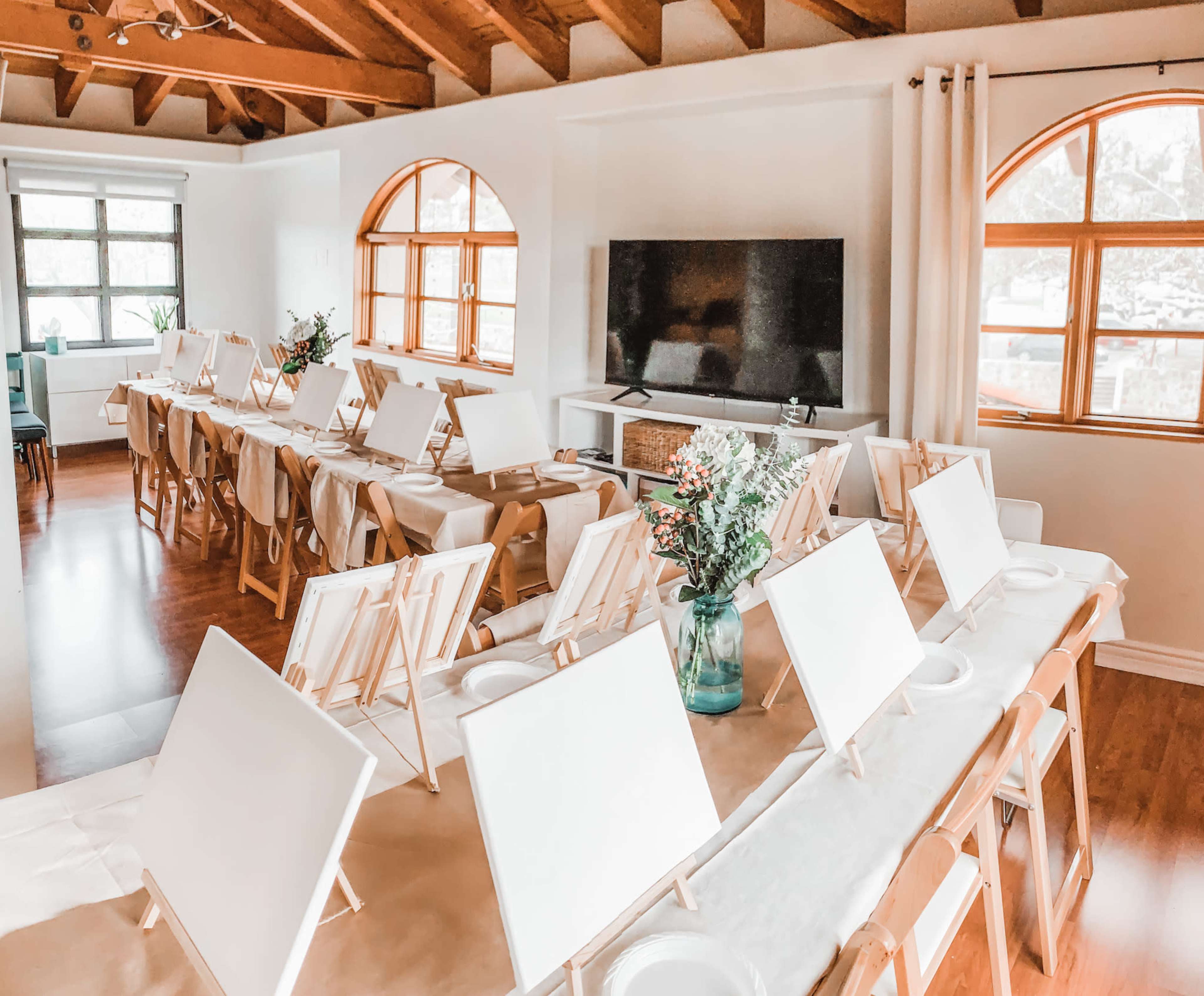 A spacious art studio with multiple easels and blank canvases arranged on long tables, surrounded by large windows and wooden beams.