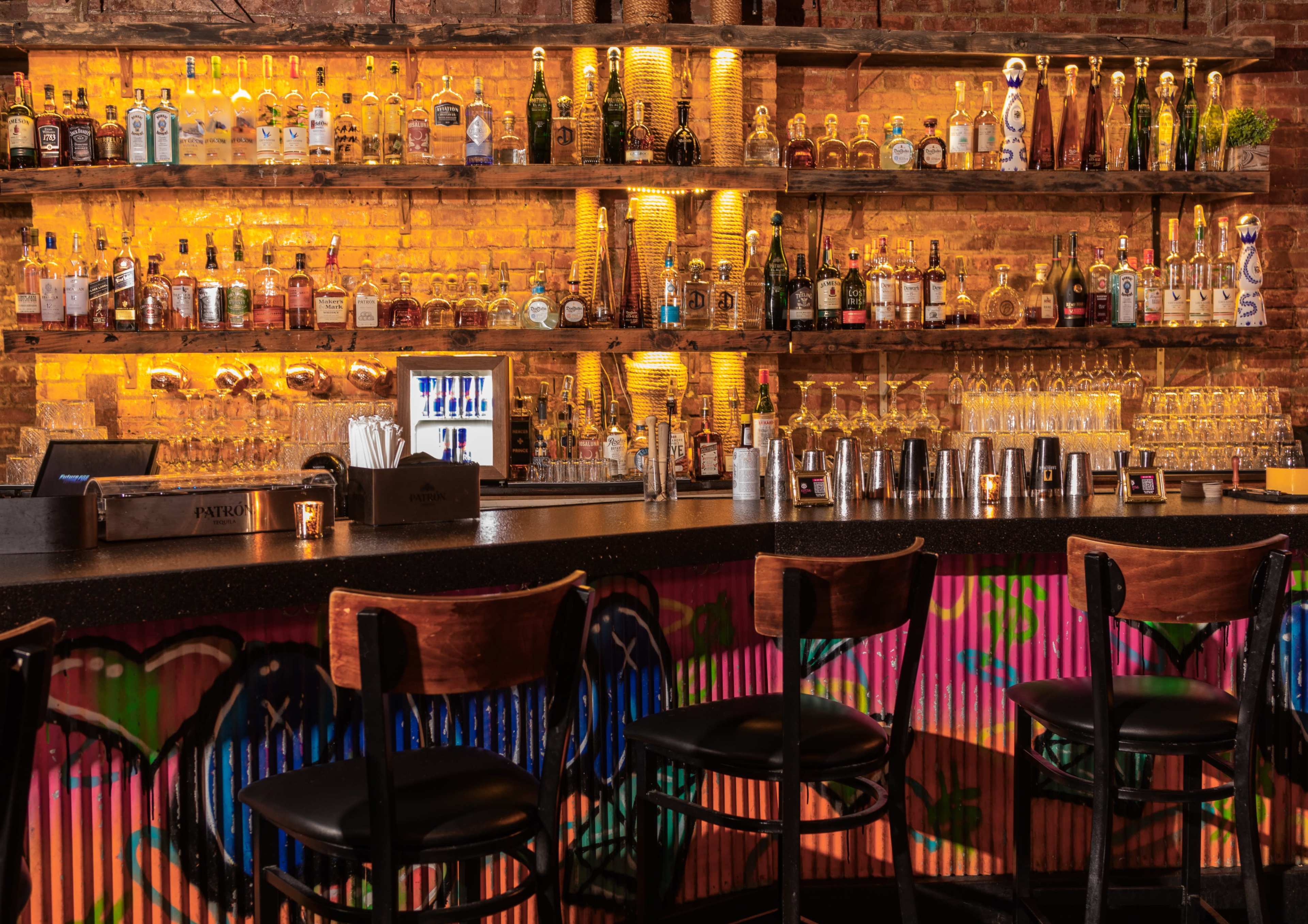 The image shows a well-stocked bar with various bottles arranged on shelves against a brick wall, illuminated by warm lighting, and empty bar stools in front.