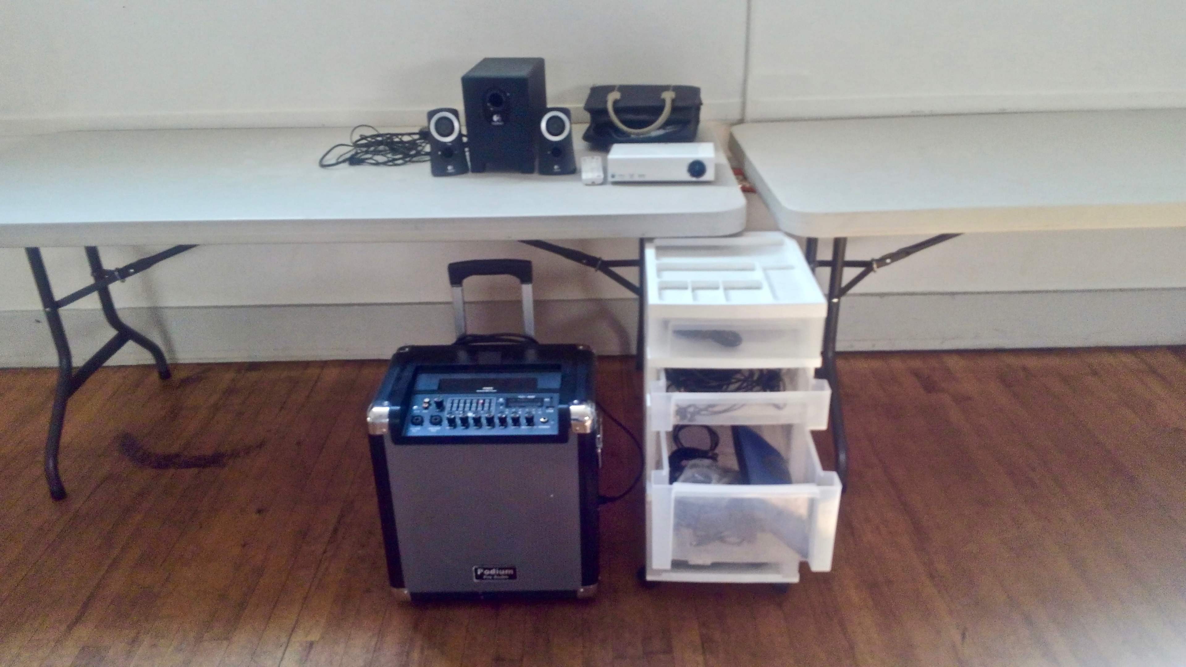 A portable sound system is placed next to a set of computer speakers and a storage unit on two folding tables.