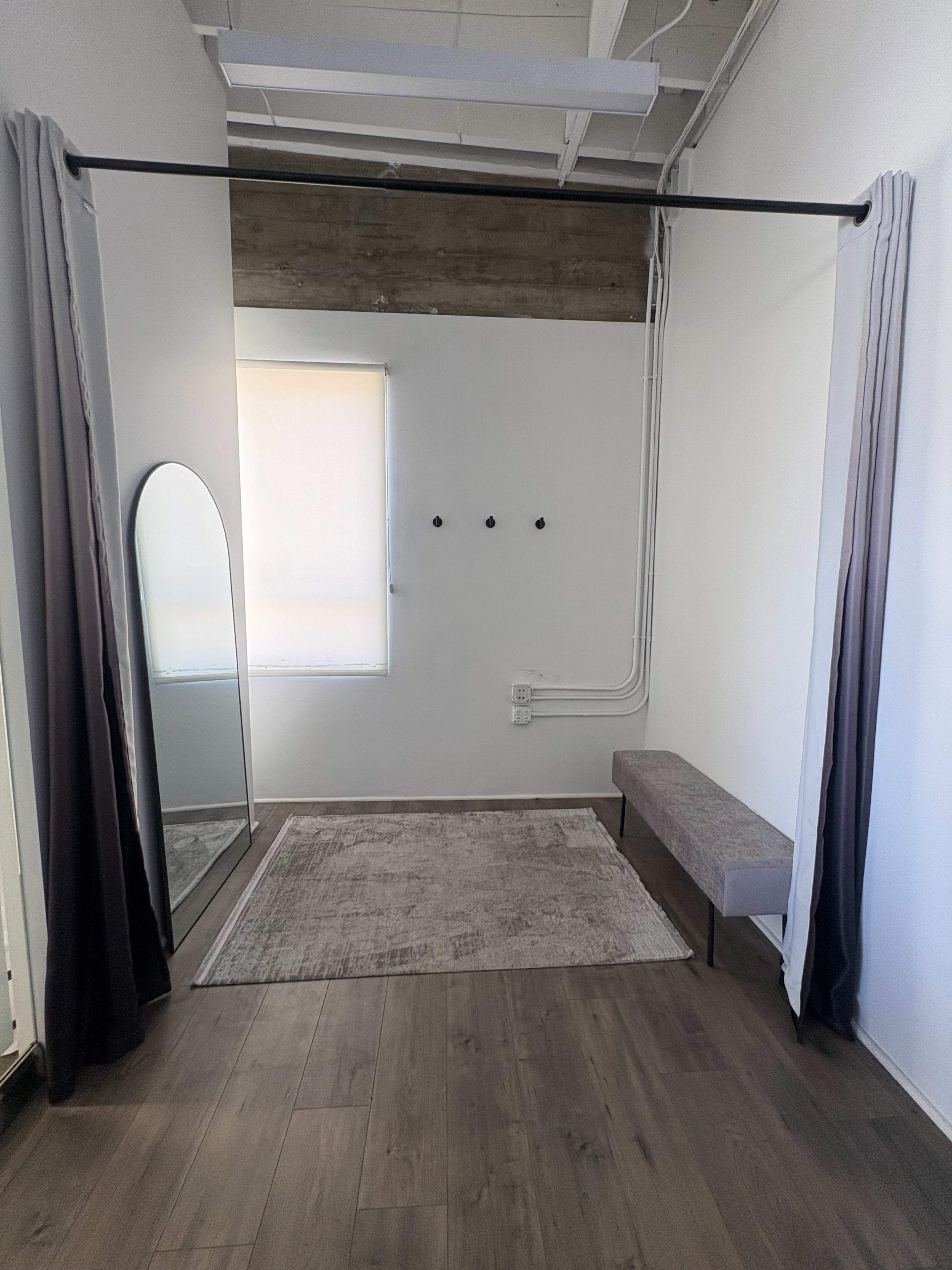 The image shows a minimalistic room featuring a large mirror, a bench, and a curtain rod with drapes, along with a rug on the floor and three wall-mounted hooks.