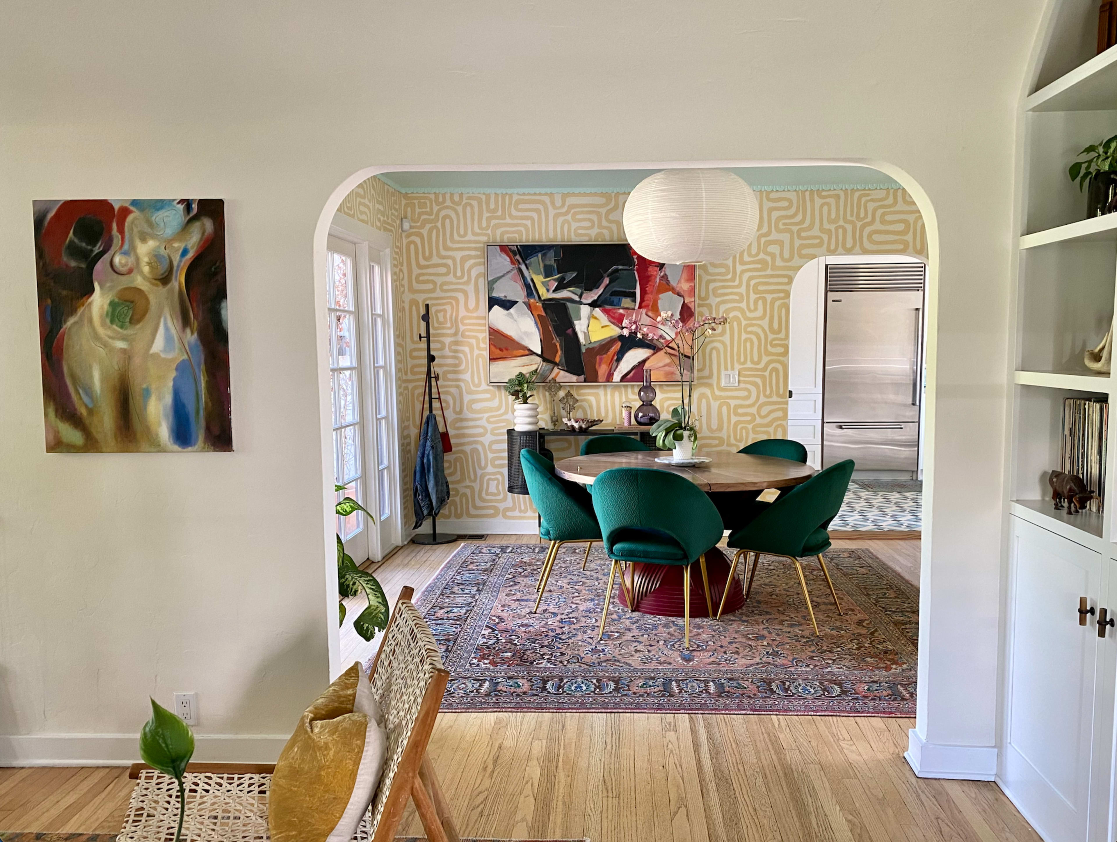 A dining area features a round table surrounded by green chairs, with abstract art on the walls, set against a patterned wallpaper and a decorative rug.