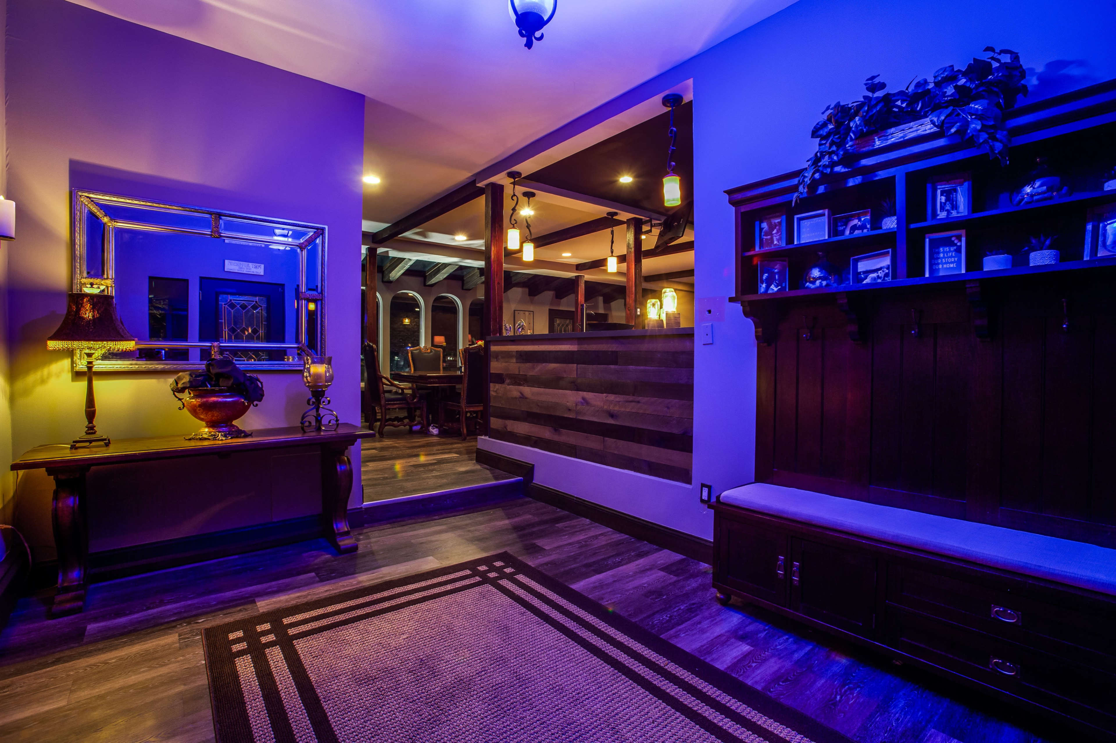 A hallway features a mirror and decorative lamp beside a bench, illuminated by blue lighting, with a dining area visible in the background.