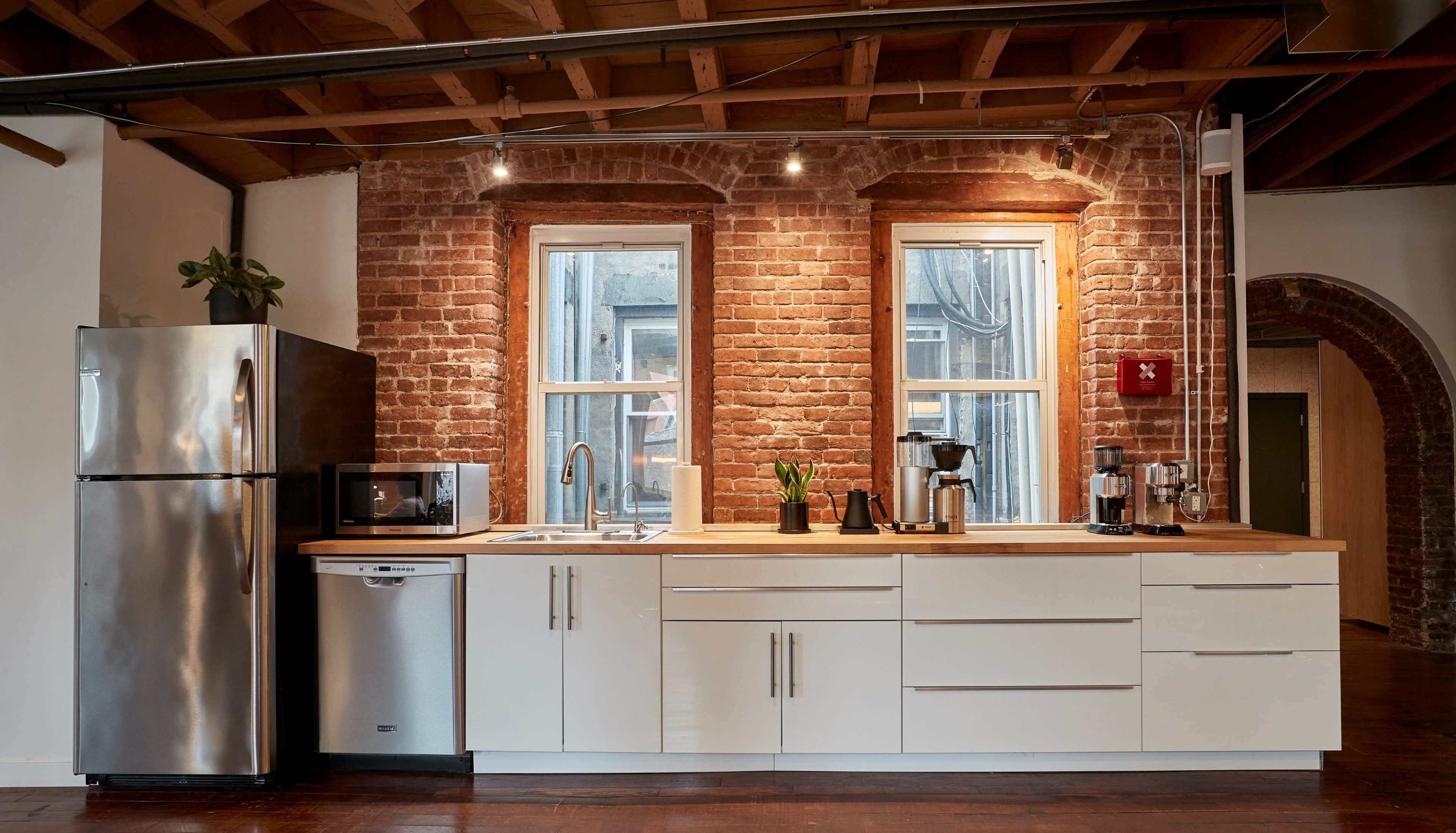 A modern kitchen with stainless steel appliances, white cabinetry, and exposed brick walls under wooden beams.