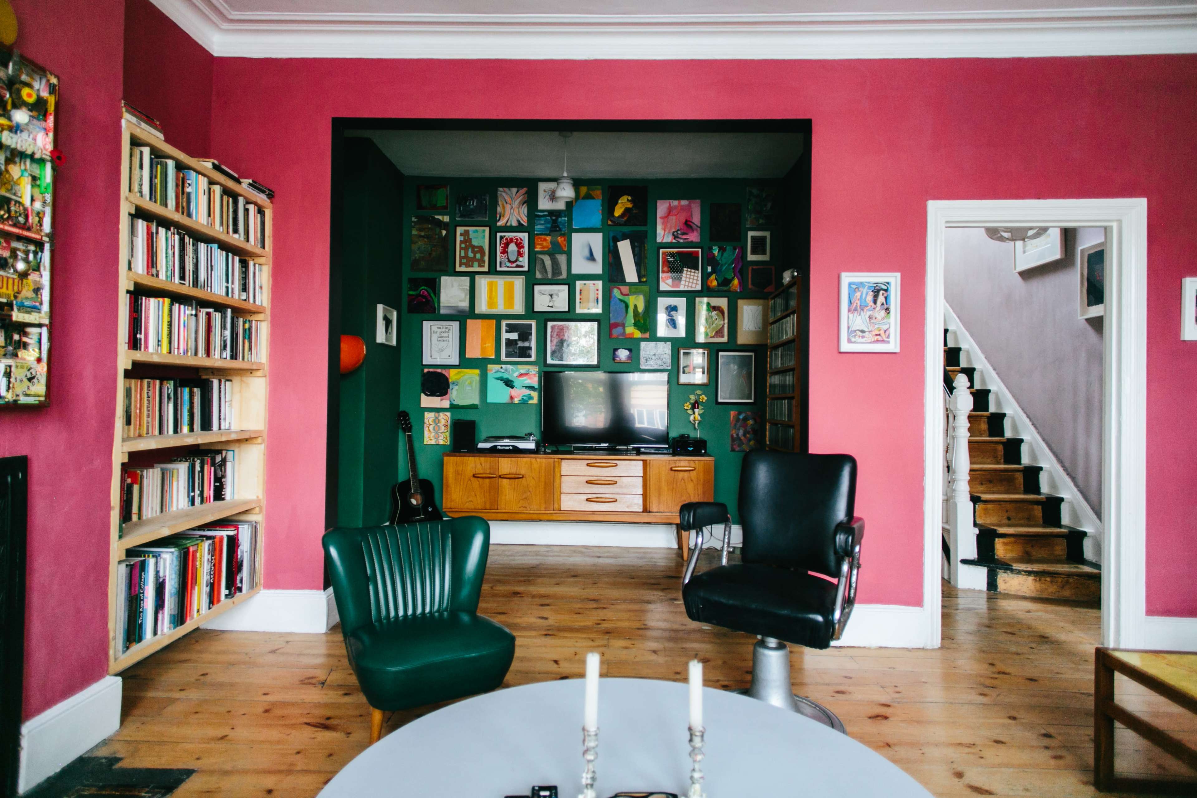 A living room features a deep pink wall, a collage of framed artworks, a wooden television cabinet, and various seating options.