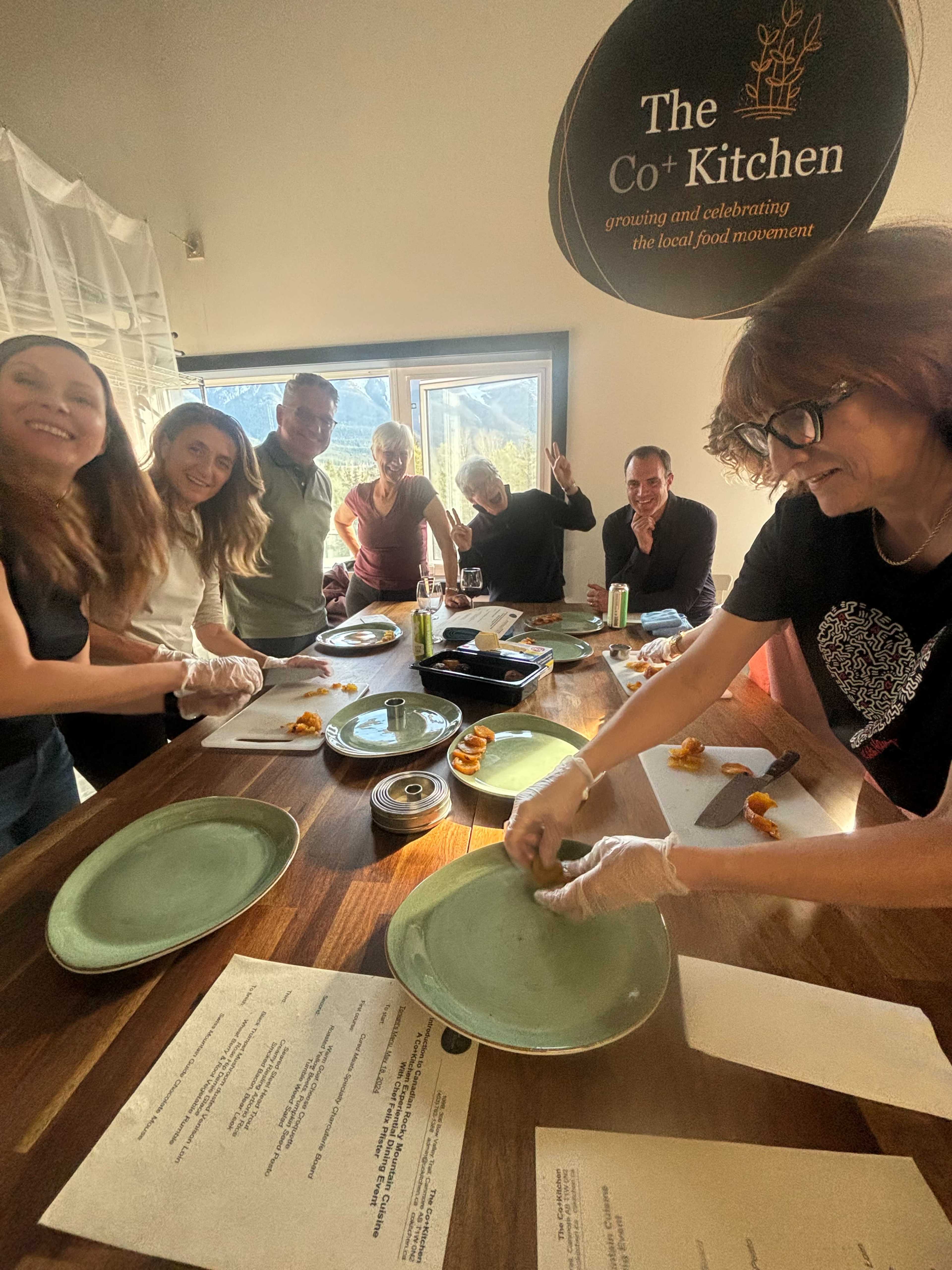 A group of six people in a kitchen gathers around a wooden table, preparing plates of food while wearing gloves.