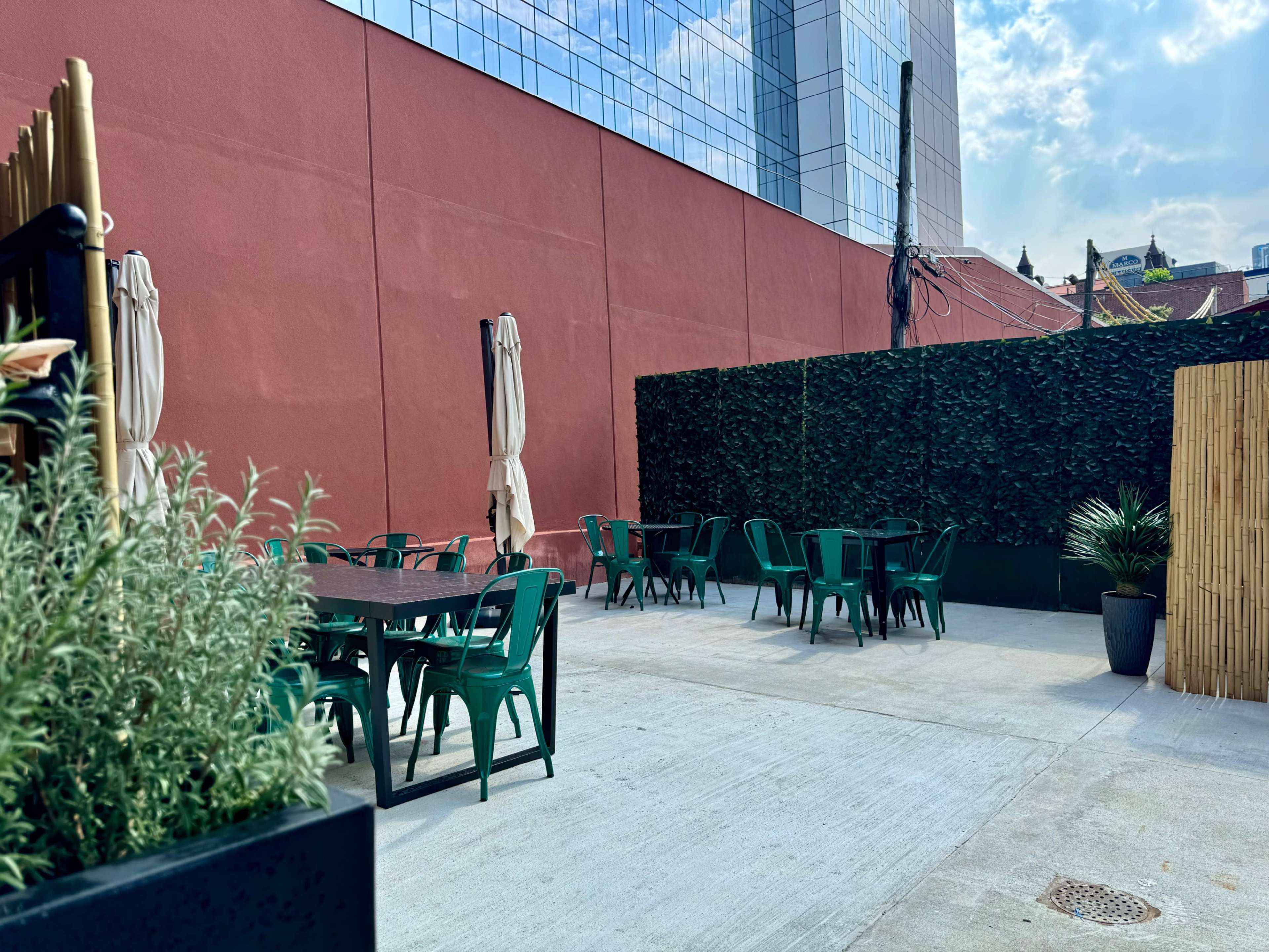 An outdoor seating area features green tables and chairs against a red wall and a backdrop of tall glass buildings.