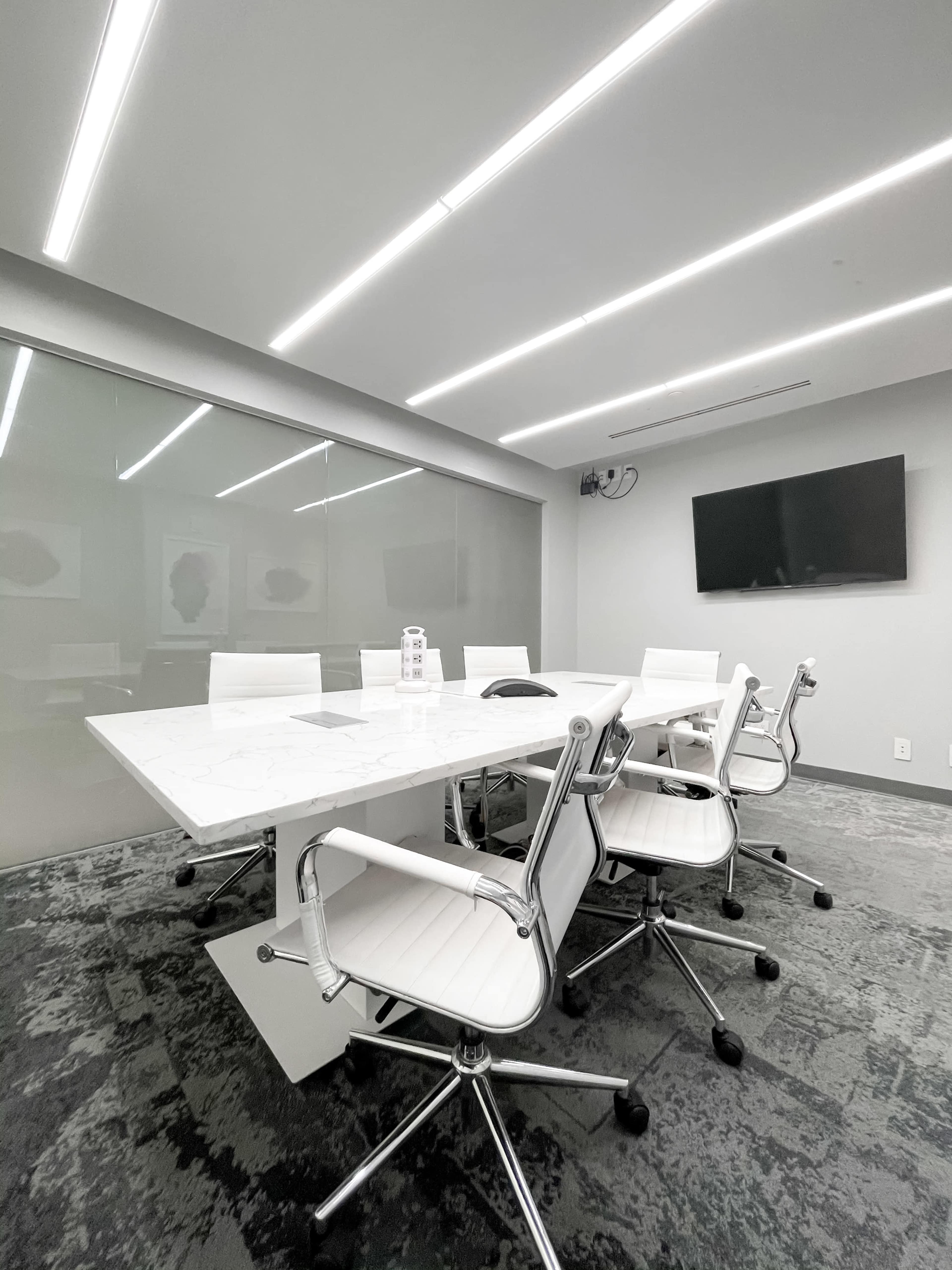 A modern conference room features a large white table surrounded by several chrome and white chairs, with a television mounted on the wall and overhead lighting.