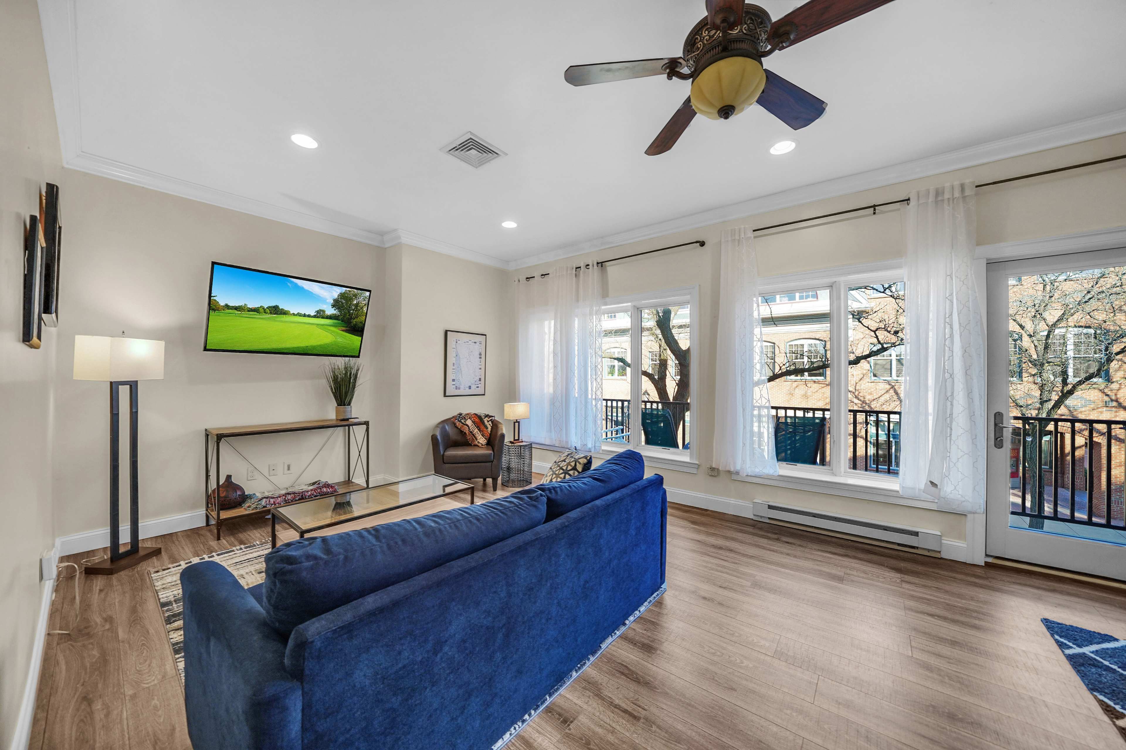 The living room features a blue sofa, a coffee table, and a television mounted on the wall, with large windows providing natural light.