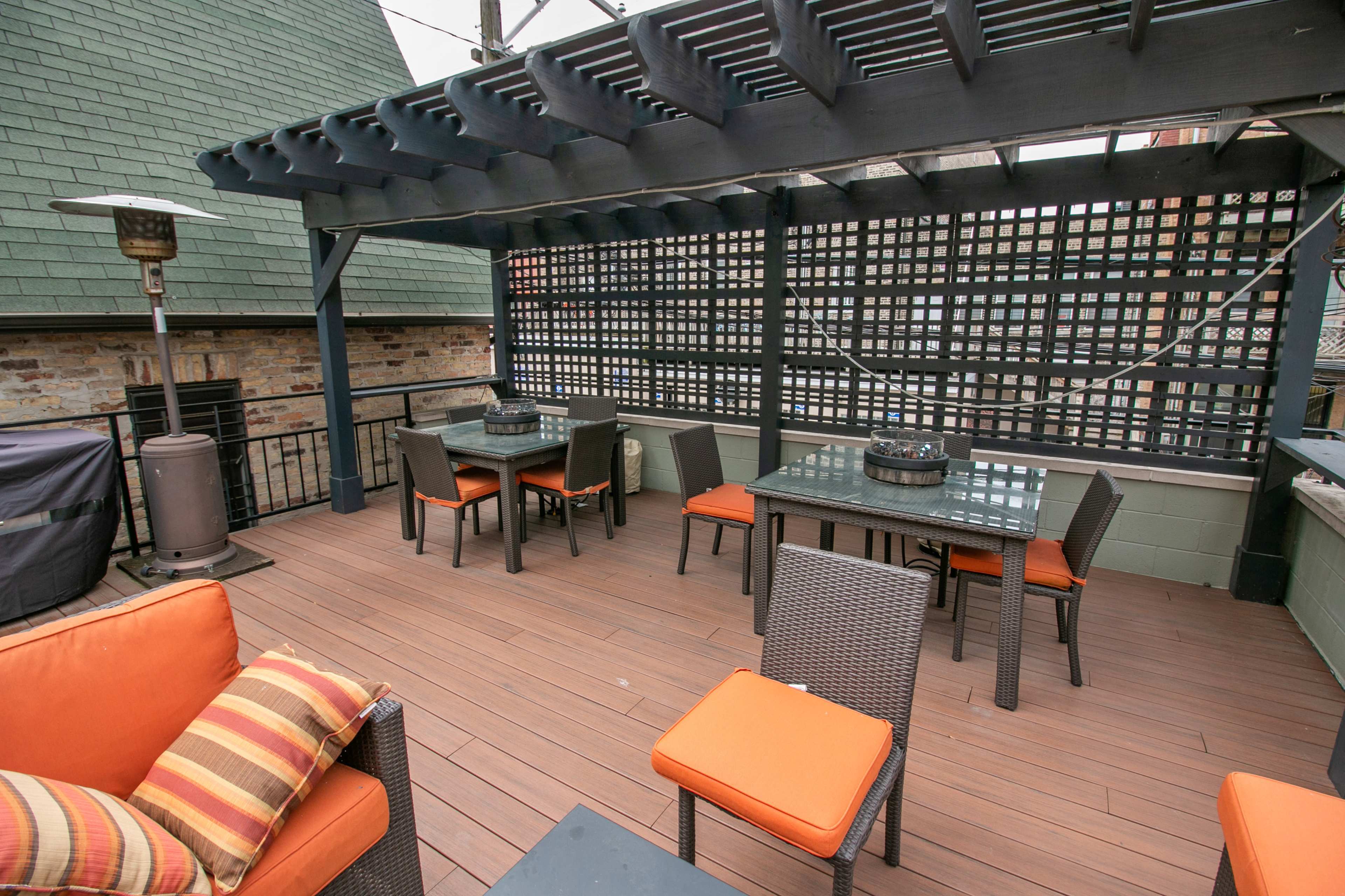 The image shows a rooftop terrace featuring a wooden deck, a pergola overhead, and several tables and chairs arranged for outdoor dining.