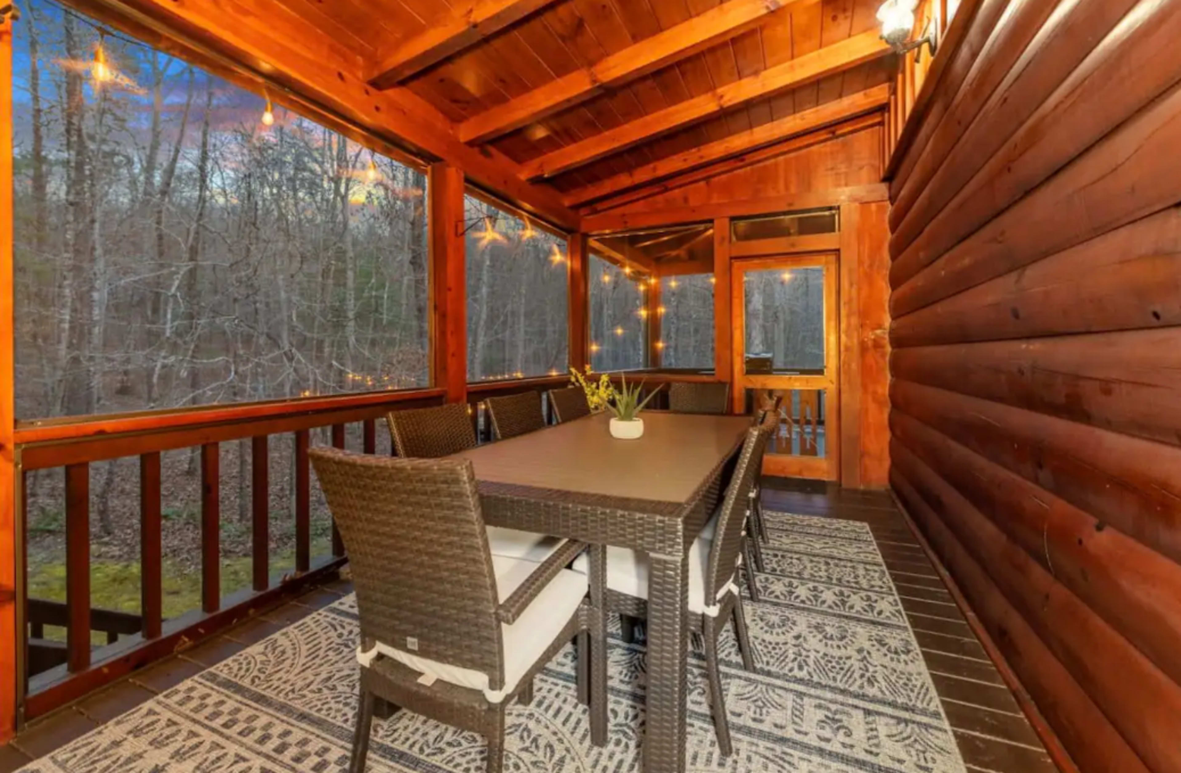 A screened-in porch features a large dining table surrounded by wicker chairs, with wooden walls and a view of trees outside.
