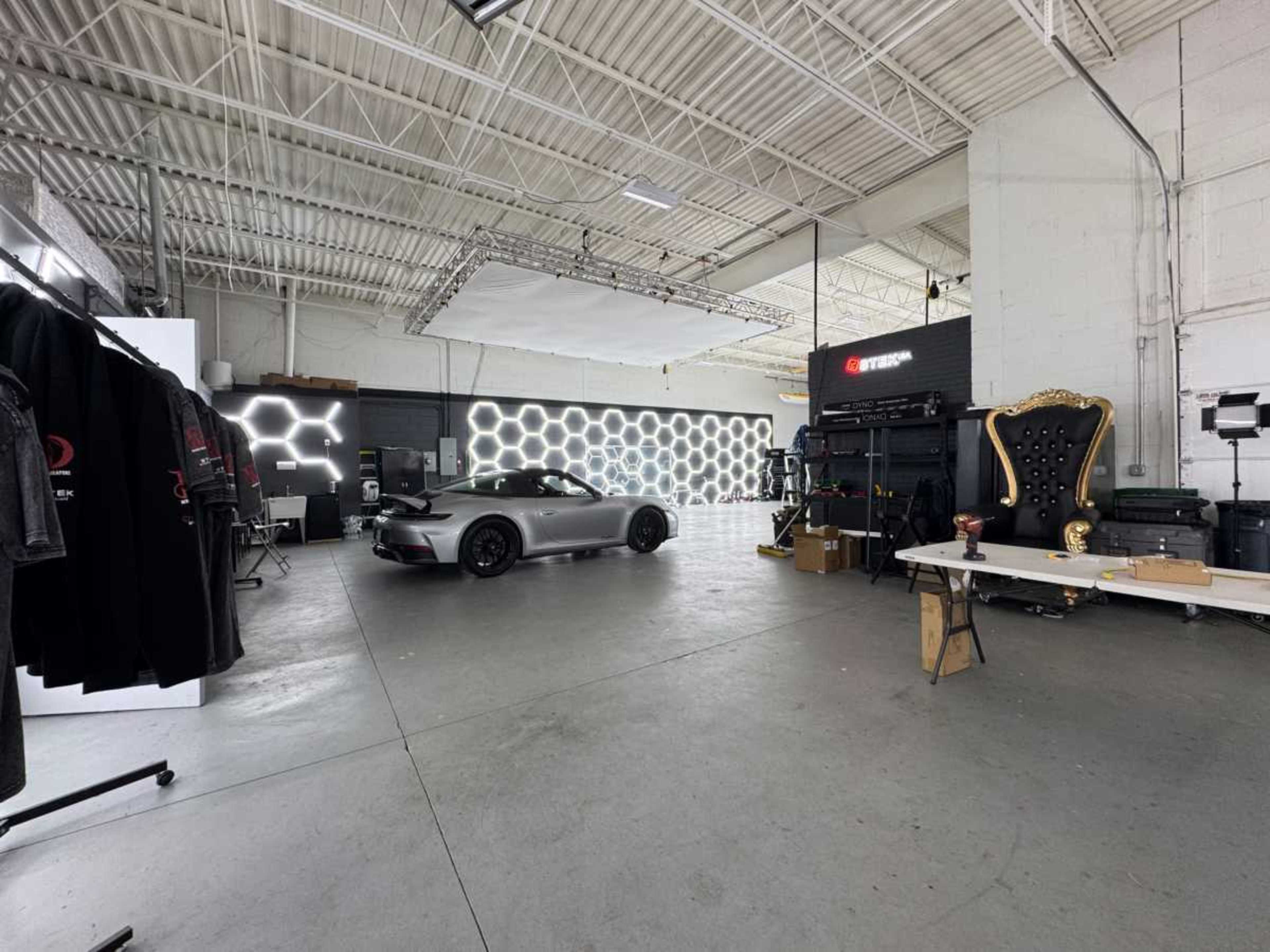 The image shows a spacious garage or studio with a sleek car parked prominently, surrounded by various equipment, clothing displays, and modern lighting features on the walls.