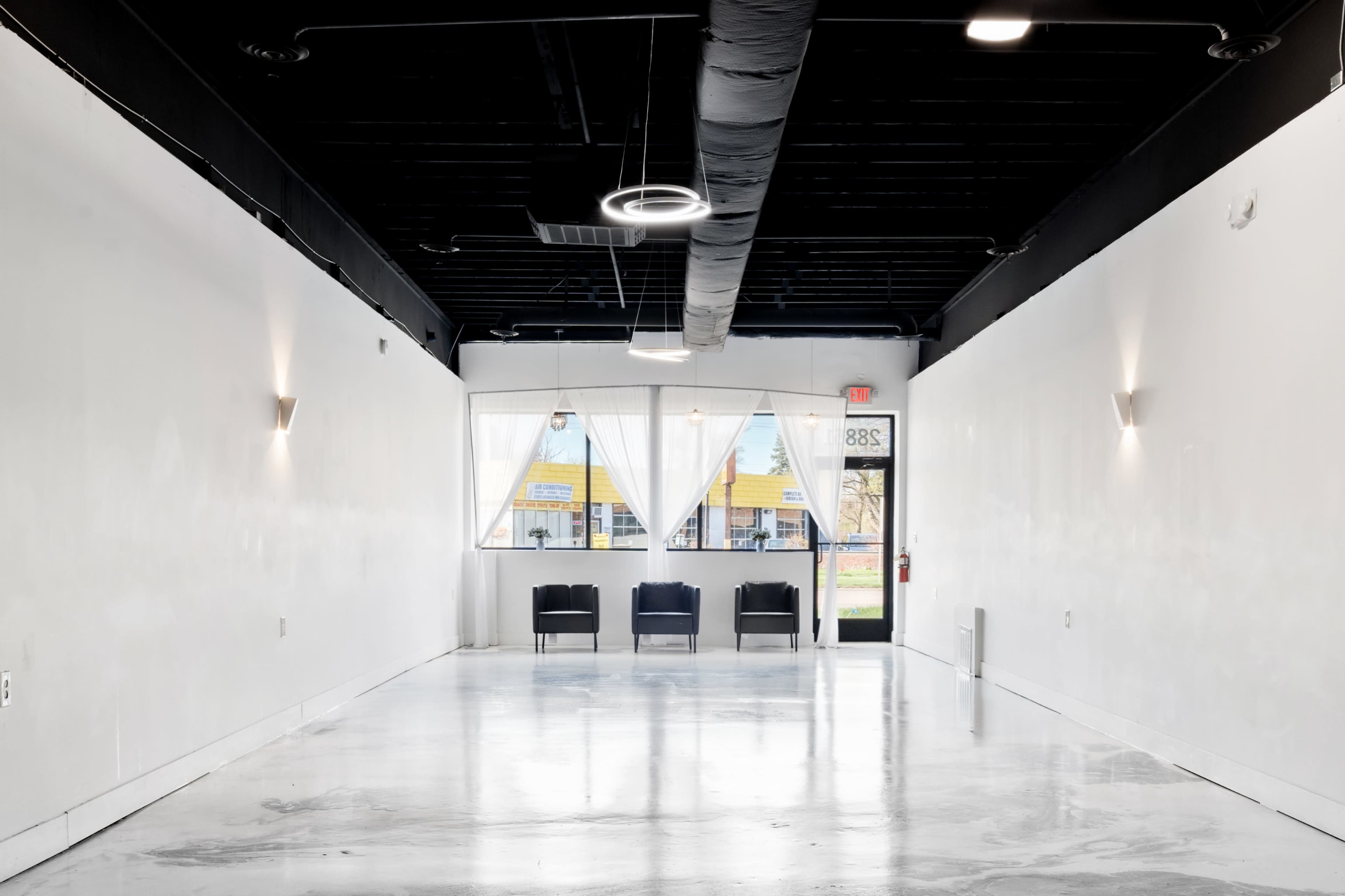 The image shows a minimalist interior of a vacant room with a polished concrete floor, white walls, and a large front window that offers a view of the outside.