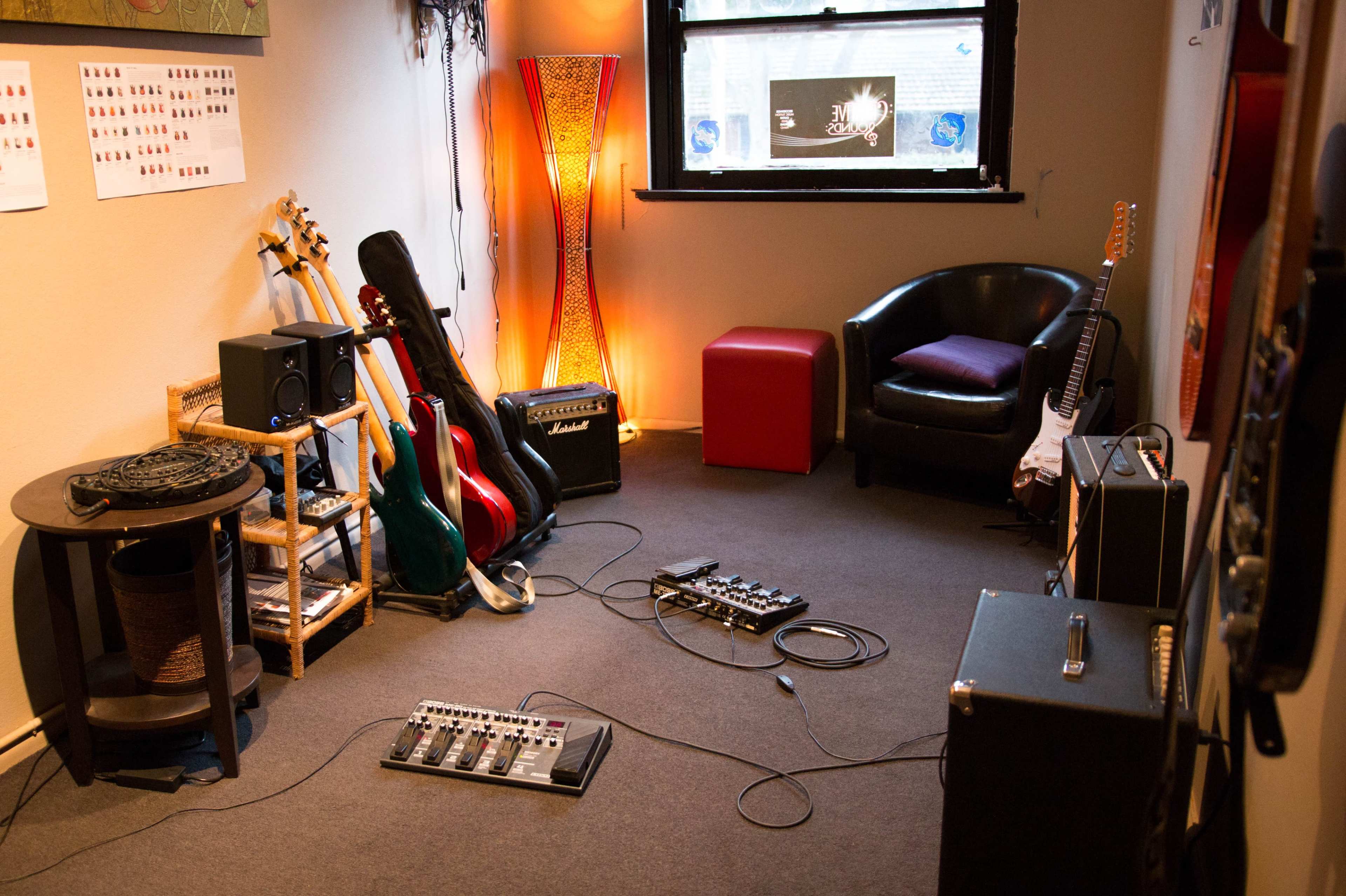 A small music practice room featuring various guitars, amplifiers, a floor pedal board, and a seating area with a chair and a small table.