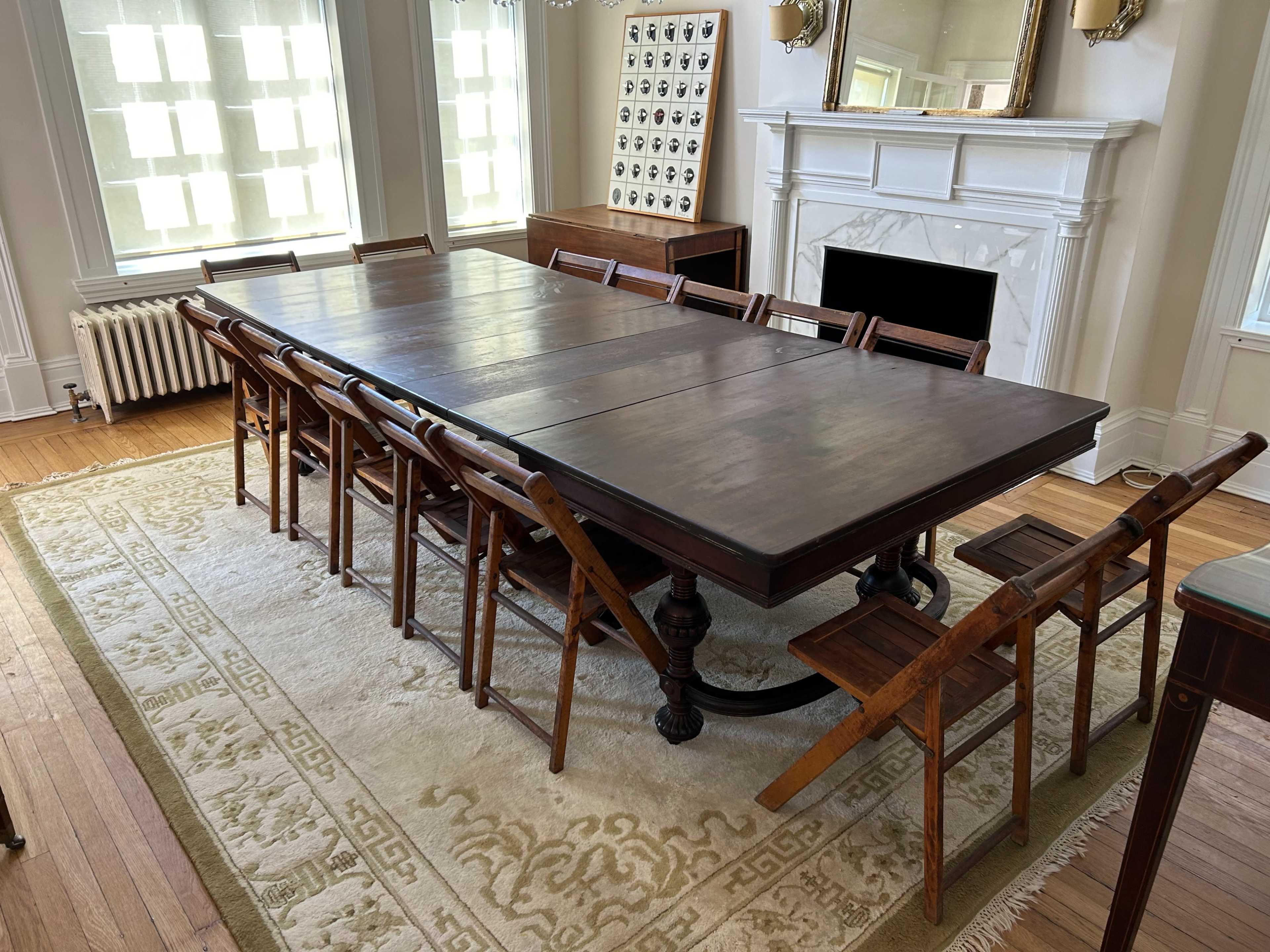 A large wooden dining table with folding chairs is set in a bright room featuring a fireplace and large windows.