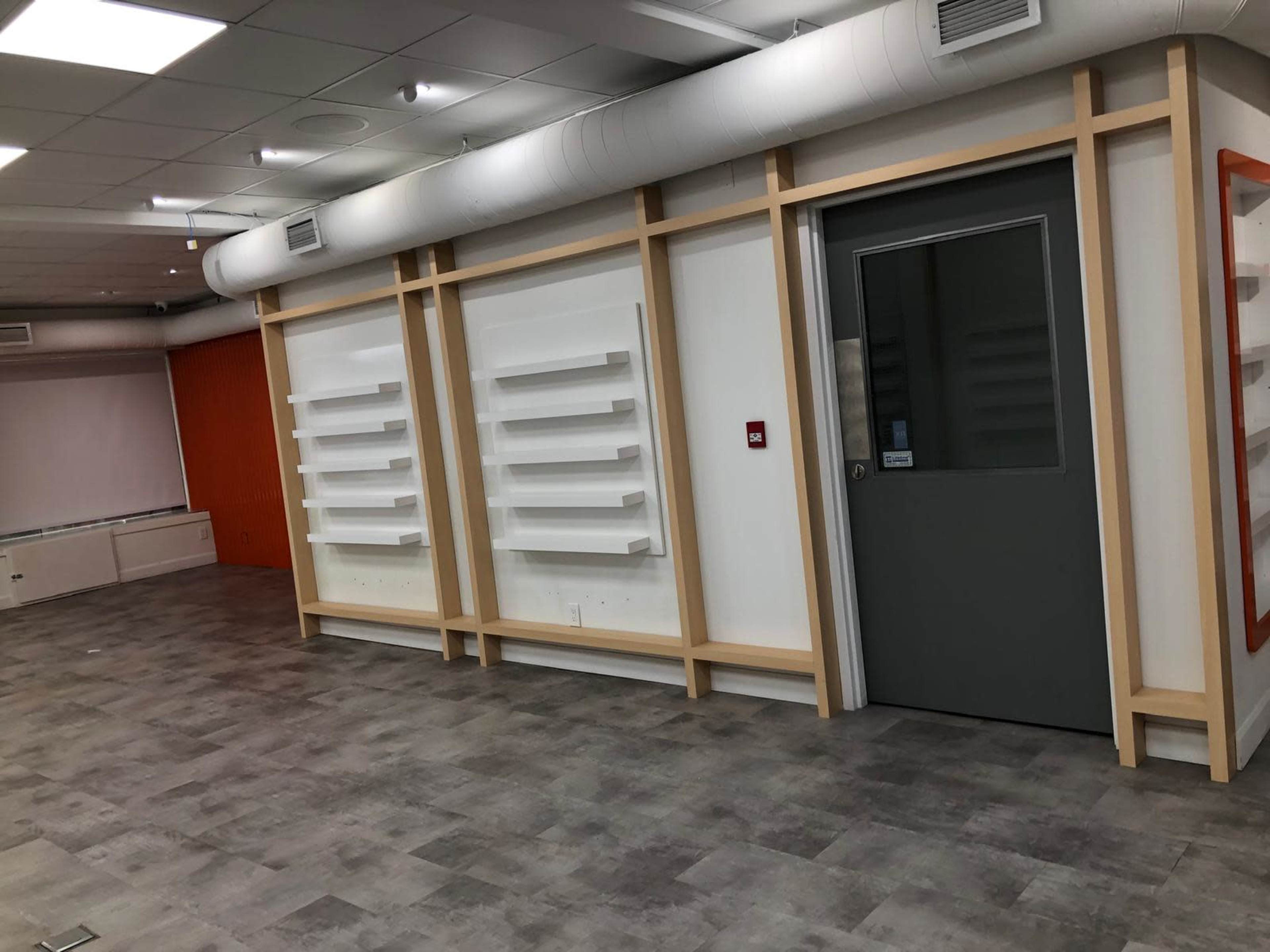 The image shows a modern, empty room featuring a doorway on the right and wall-mounted shelves along the left wall, with a gray floor and bright ceiling lights.