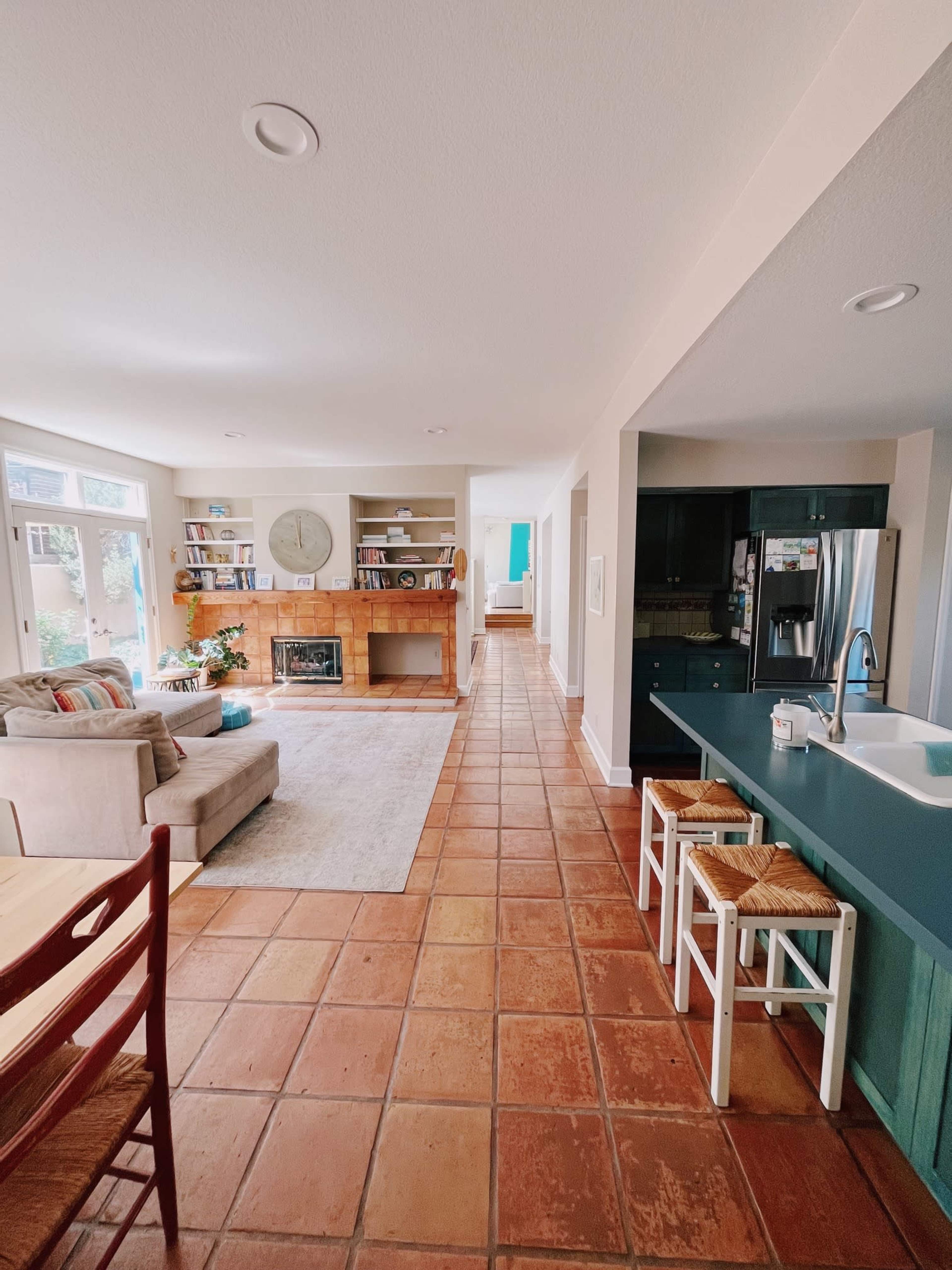The image shows a spacious living area connected to a kitchen, featuring a tiled floor, a fireplace with built-in shelves, and a hallway leading to another room.
