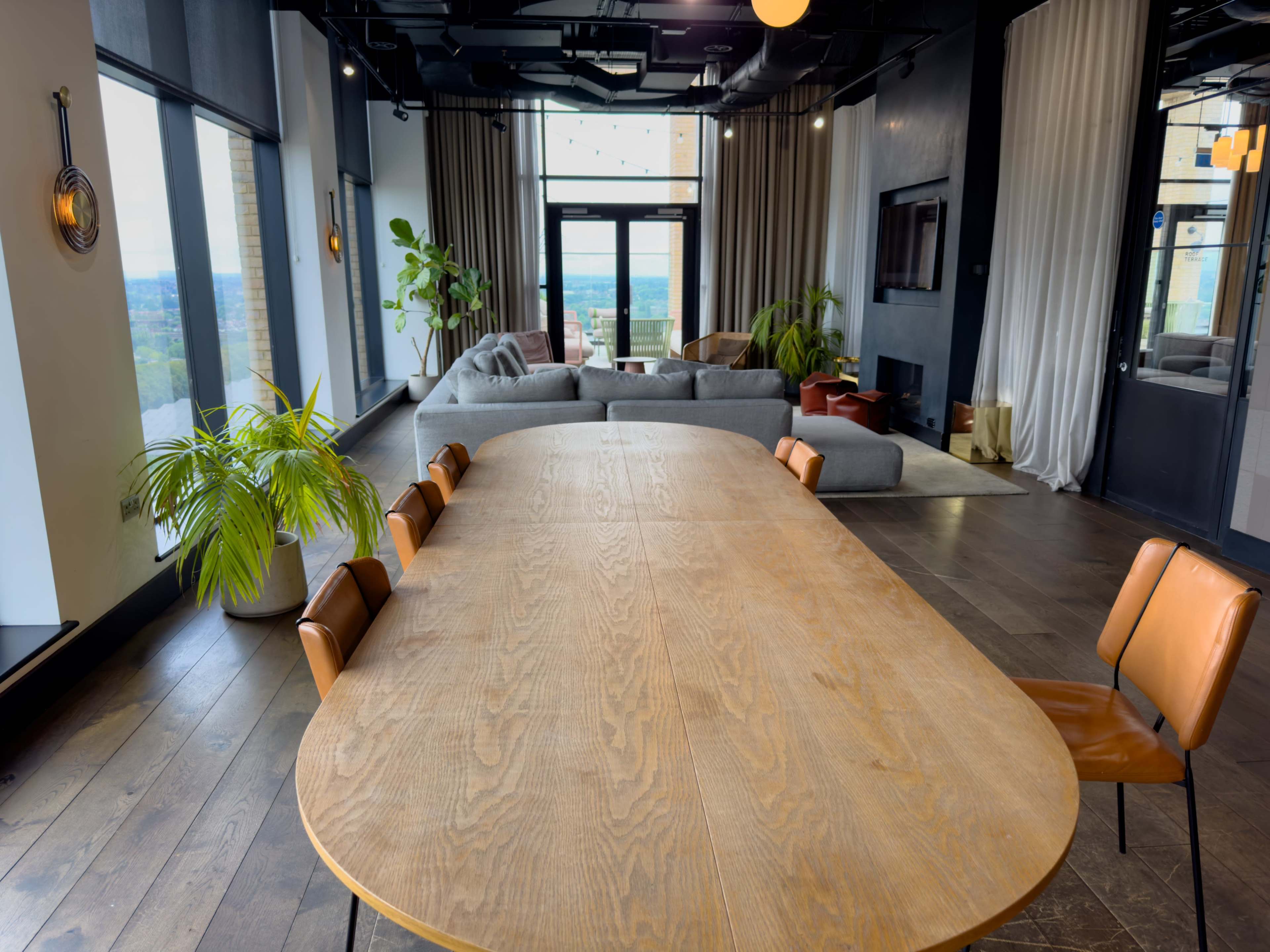 A long wooden dining table is positioned in a spacious room with large windows, overlooking a view of the outdoors and furnished with a sectional couch.