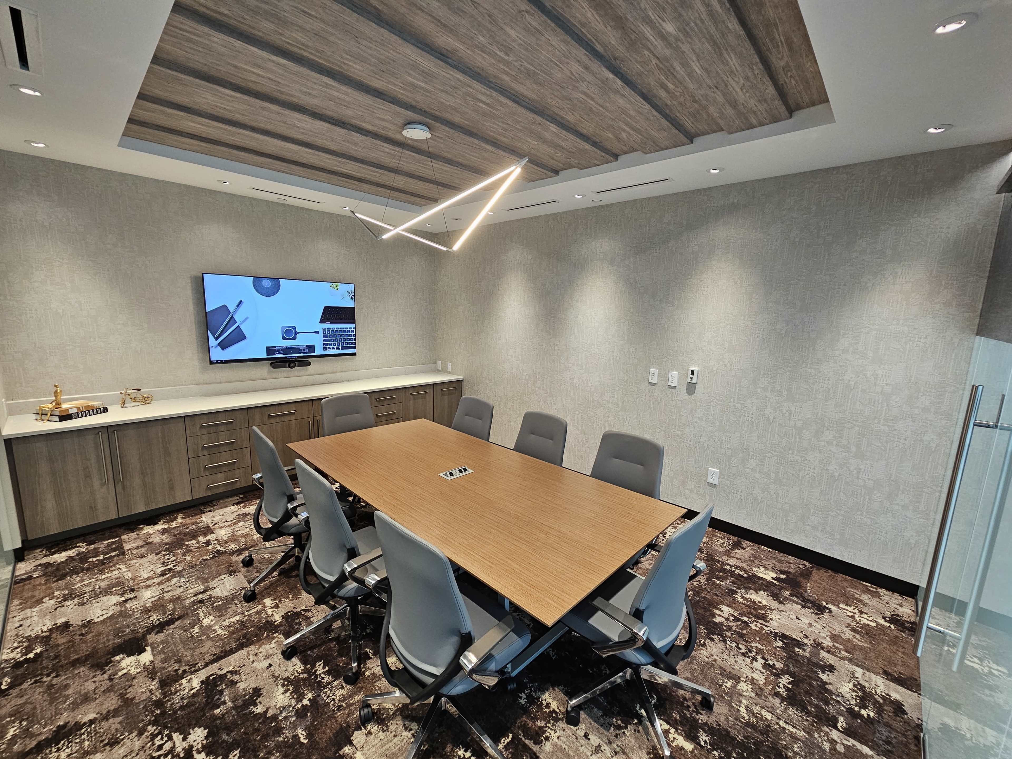 A modern conference room featuring a large wooden table surrounded by six chairs, with a television on the wall displaying a presentation.