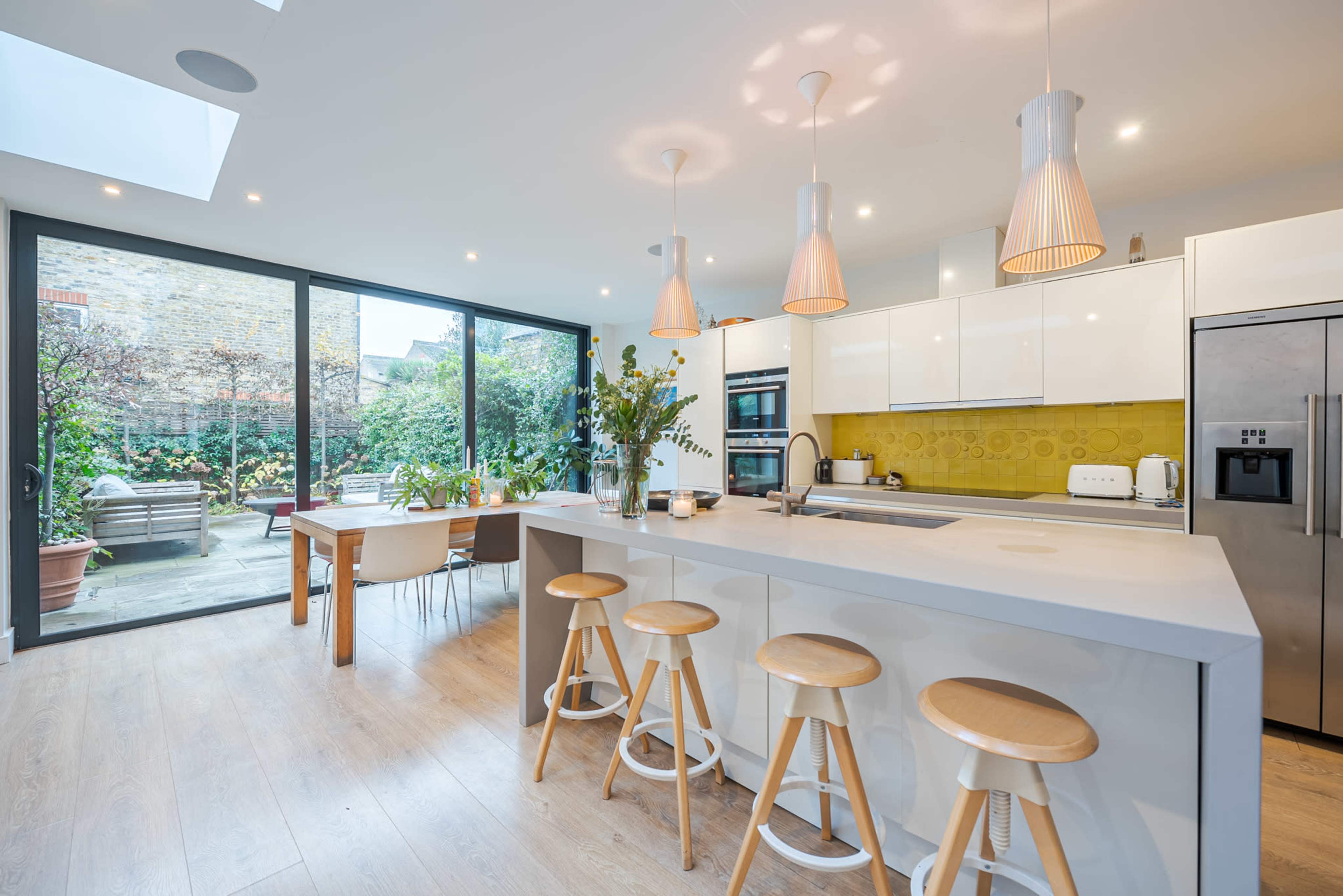 The image shows a modern kitchen with a central island, white cabinetry, pendant lights, and large glass doors leading to an outdoor garden.