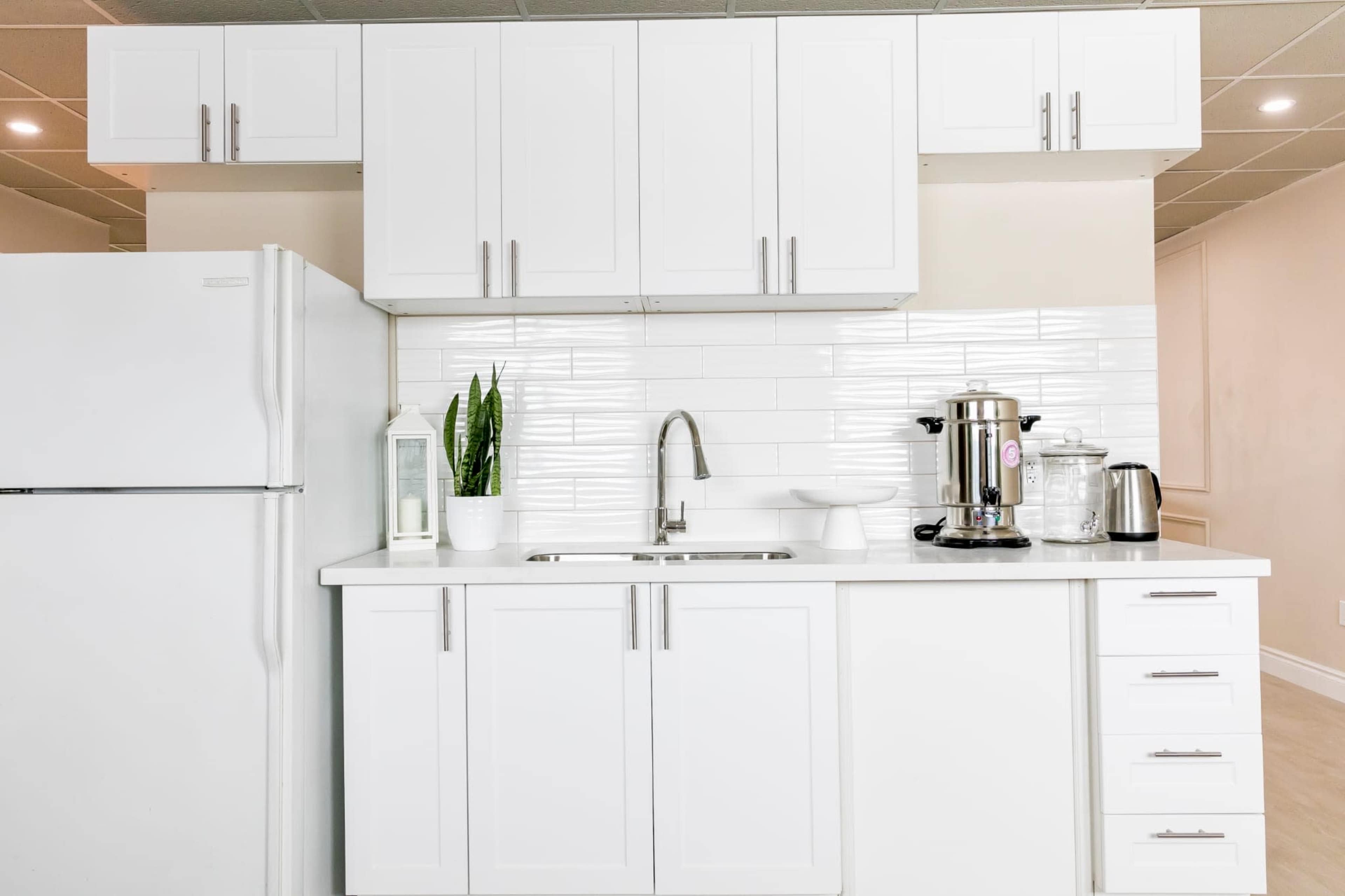 A modern kitchen with white cabinets, a stainless steel sink, and appliances including a refrigerator, coffee maker, and kettle.