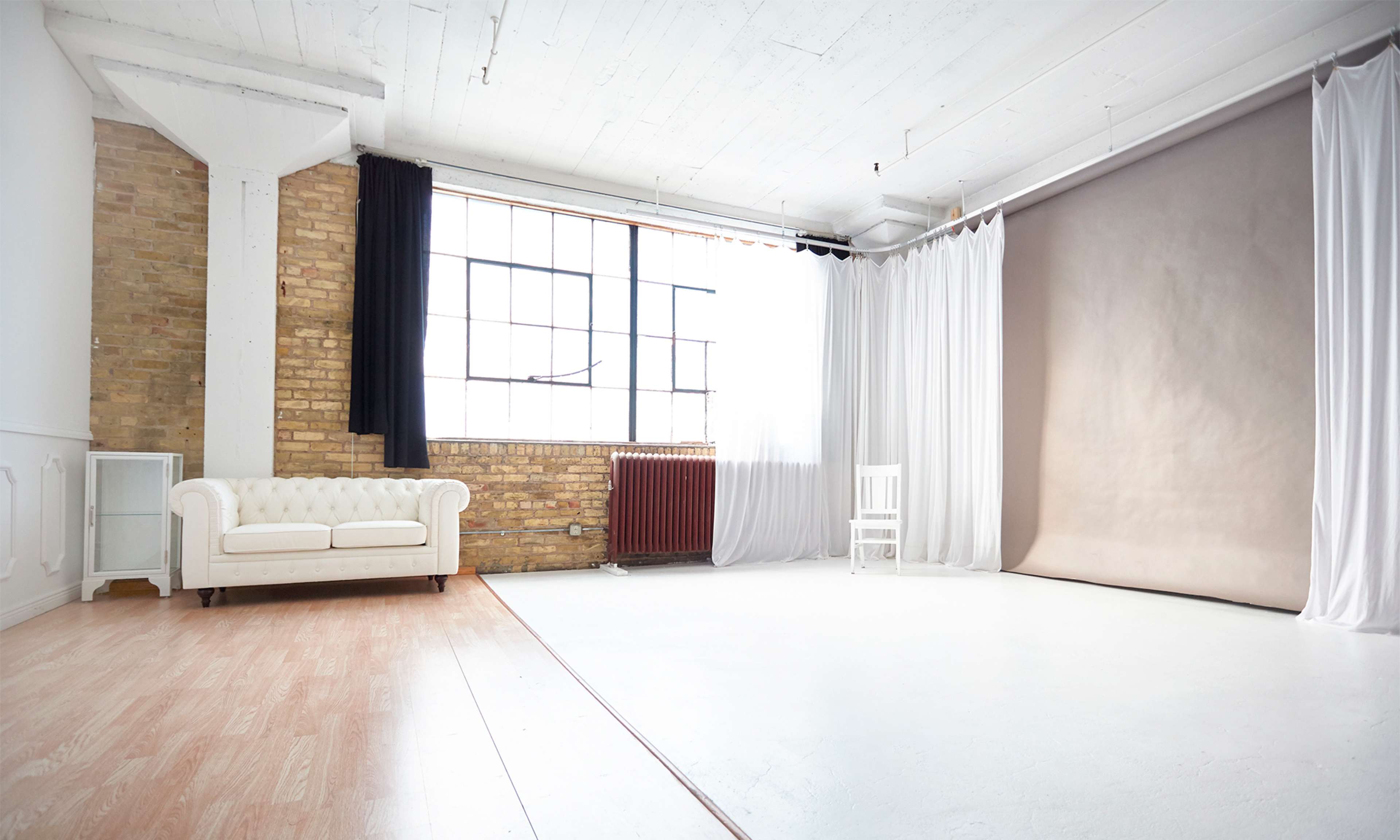 The image shows a bright, empty studio space featuring a white wooden floor, large windows, a brick wall, a white couch, and a backdrop with curtains.