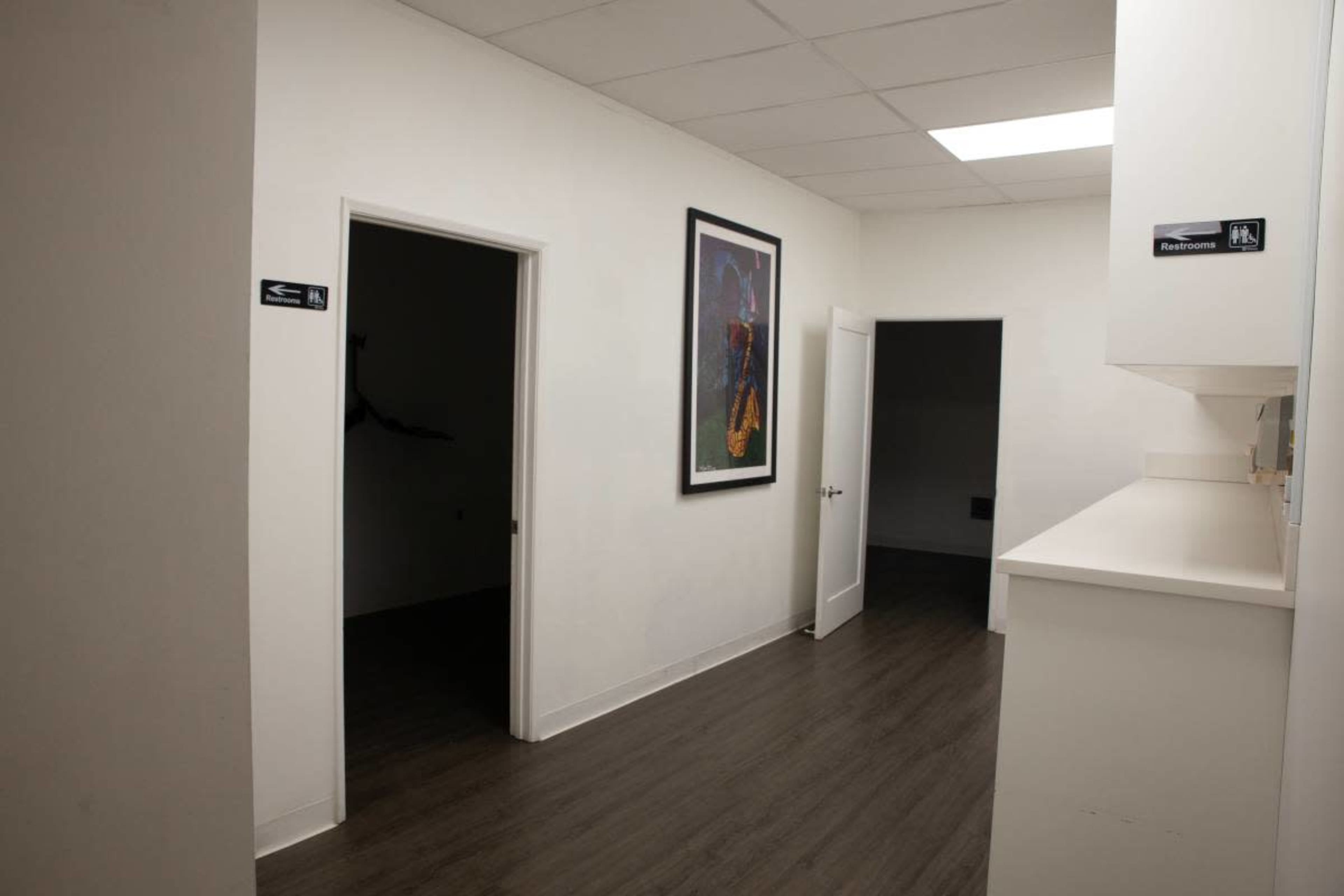 The image shows a narrow hallway with white walls, featuring two doors on the left side and a large framed artwork on the right wall.