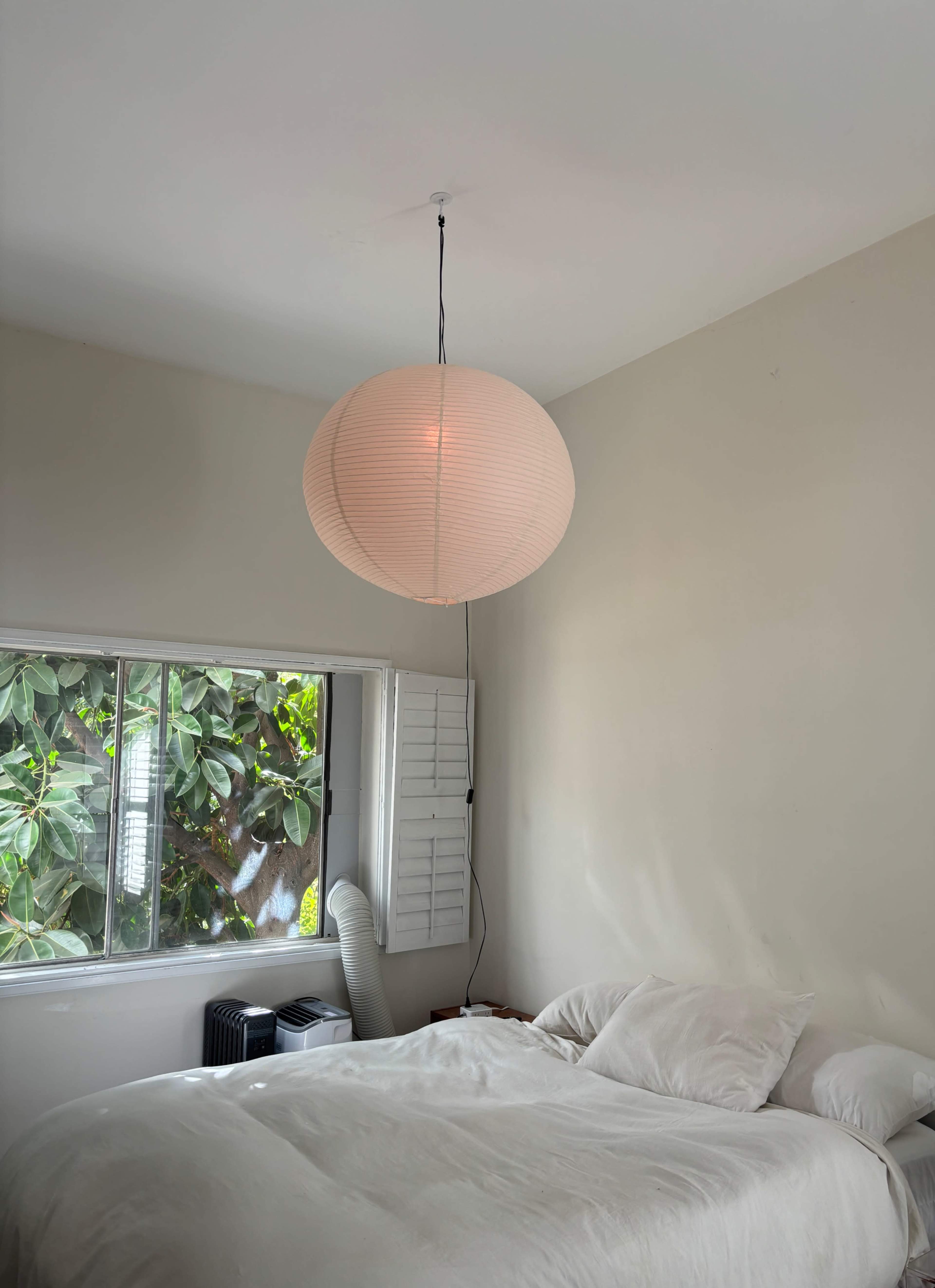 A round, white paper lantern hangs from the ceiling over a bed with white linens in a bright bedroom with a window featuring greenery outside.