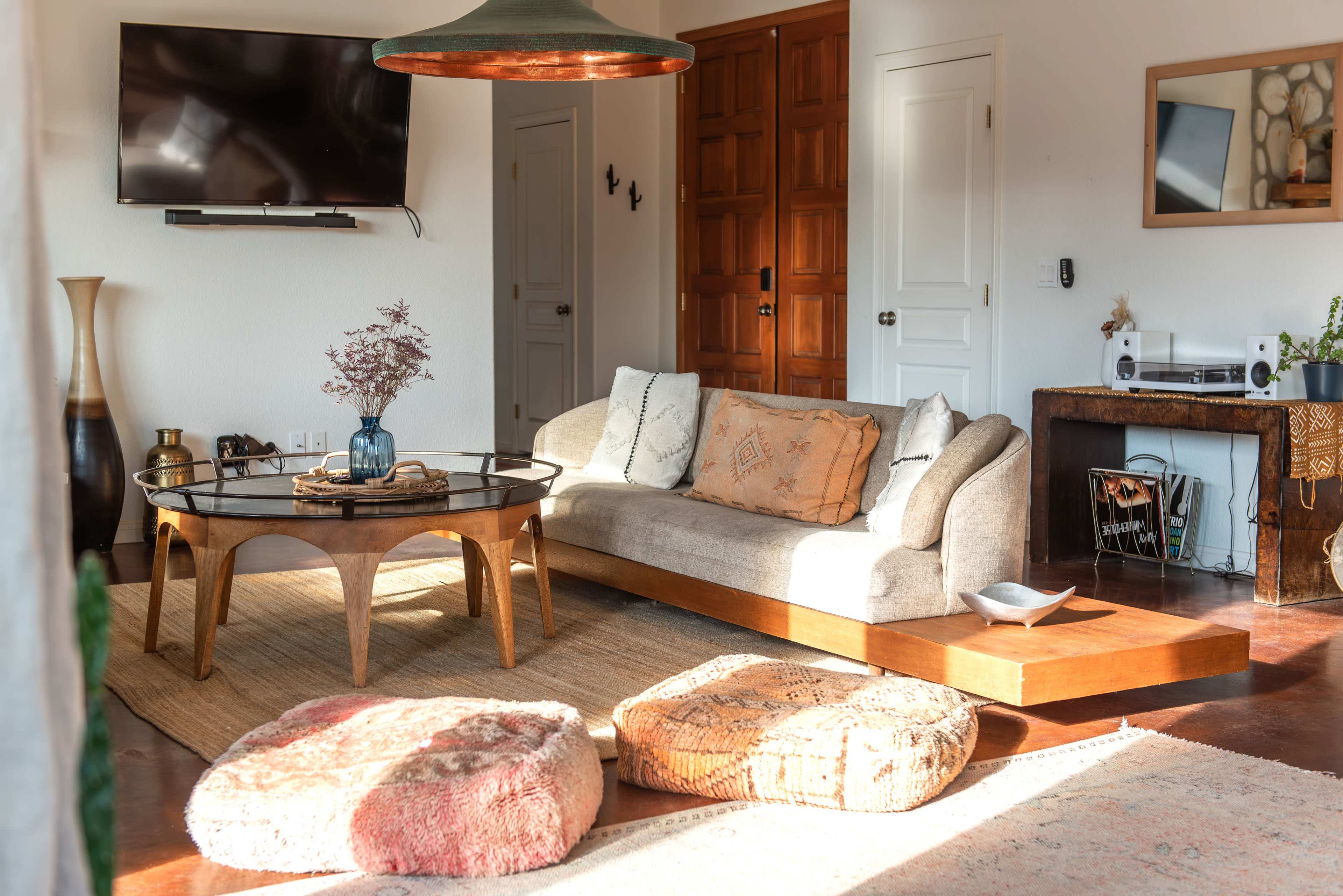 The image shows a modern living room featuring a light-colored sofa, a round coffee table, and floor cushions, with a television mounted on the wall and a wooden cabinet in the background.