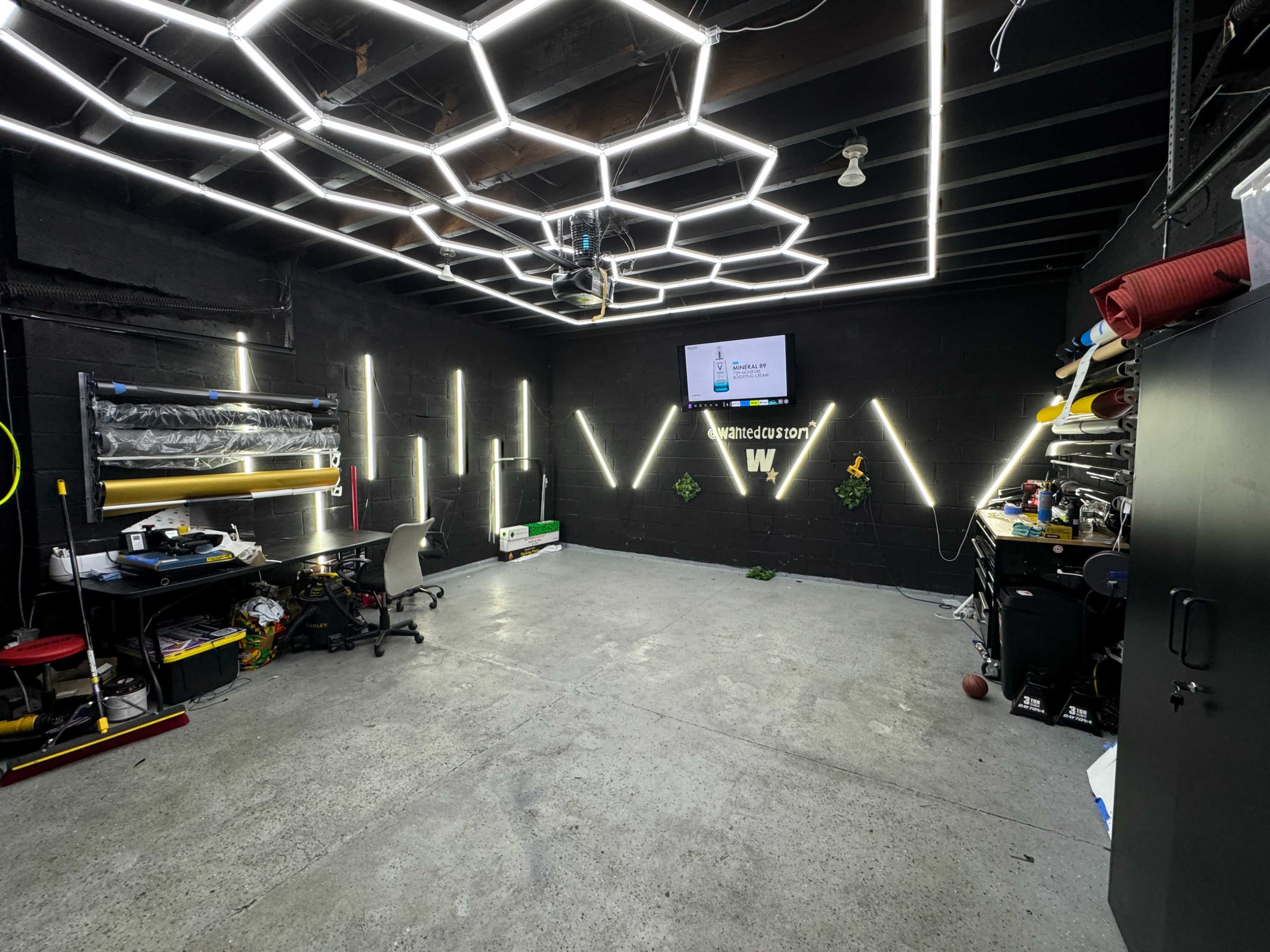 A modern garage or workshop space with hexagonal LED lighting, a television mounted on the wall, and various workstations and storage areas.