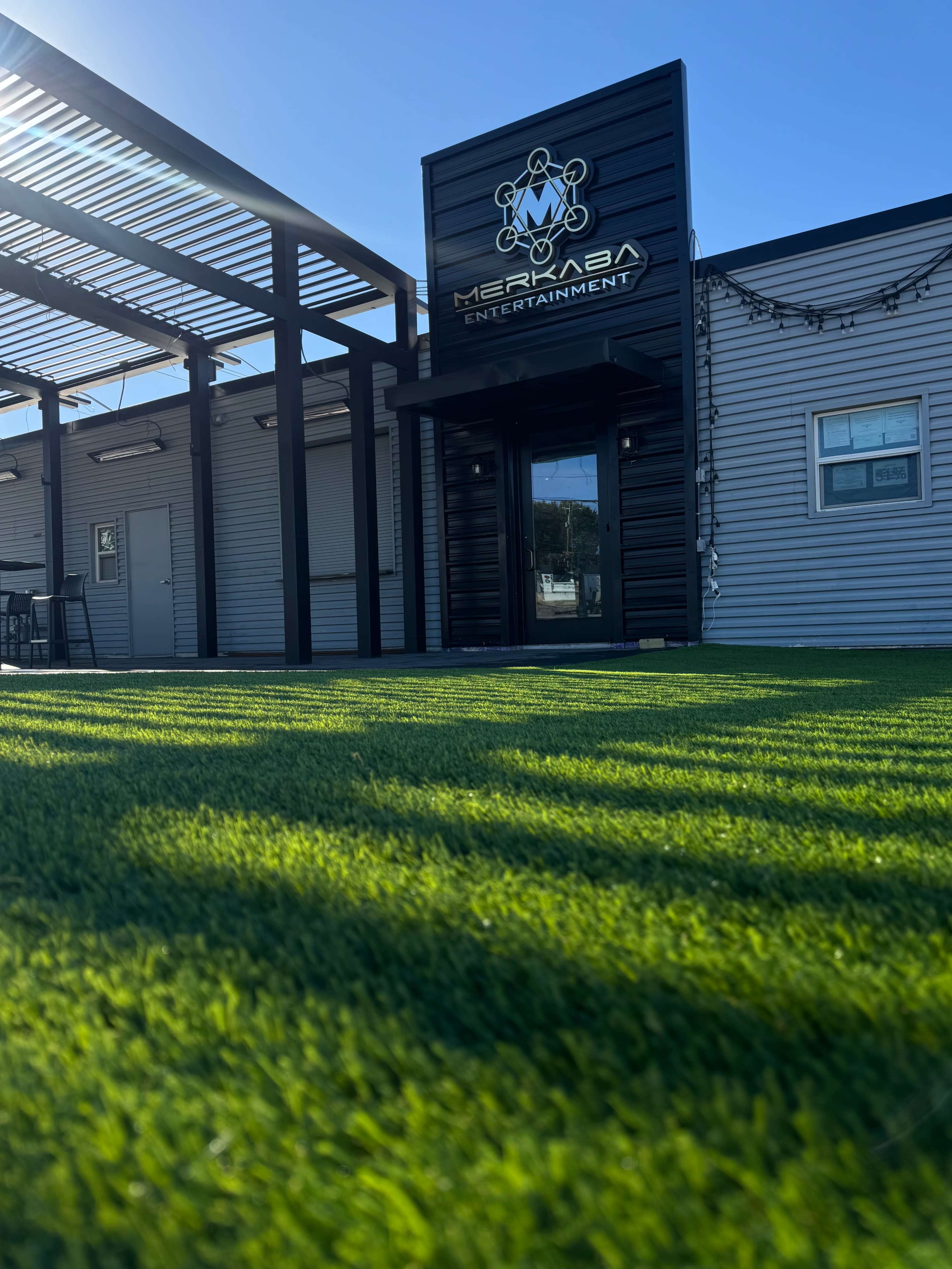 The image shows a modern building with a sign reading "Merkaba Entertainment," surrounded by a green lawn and shadowed by a pergola.