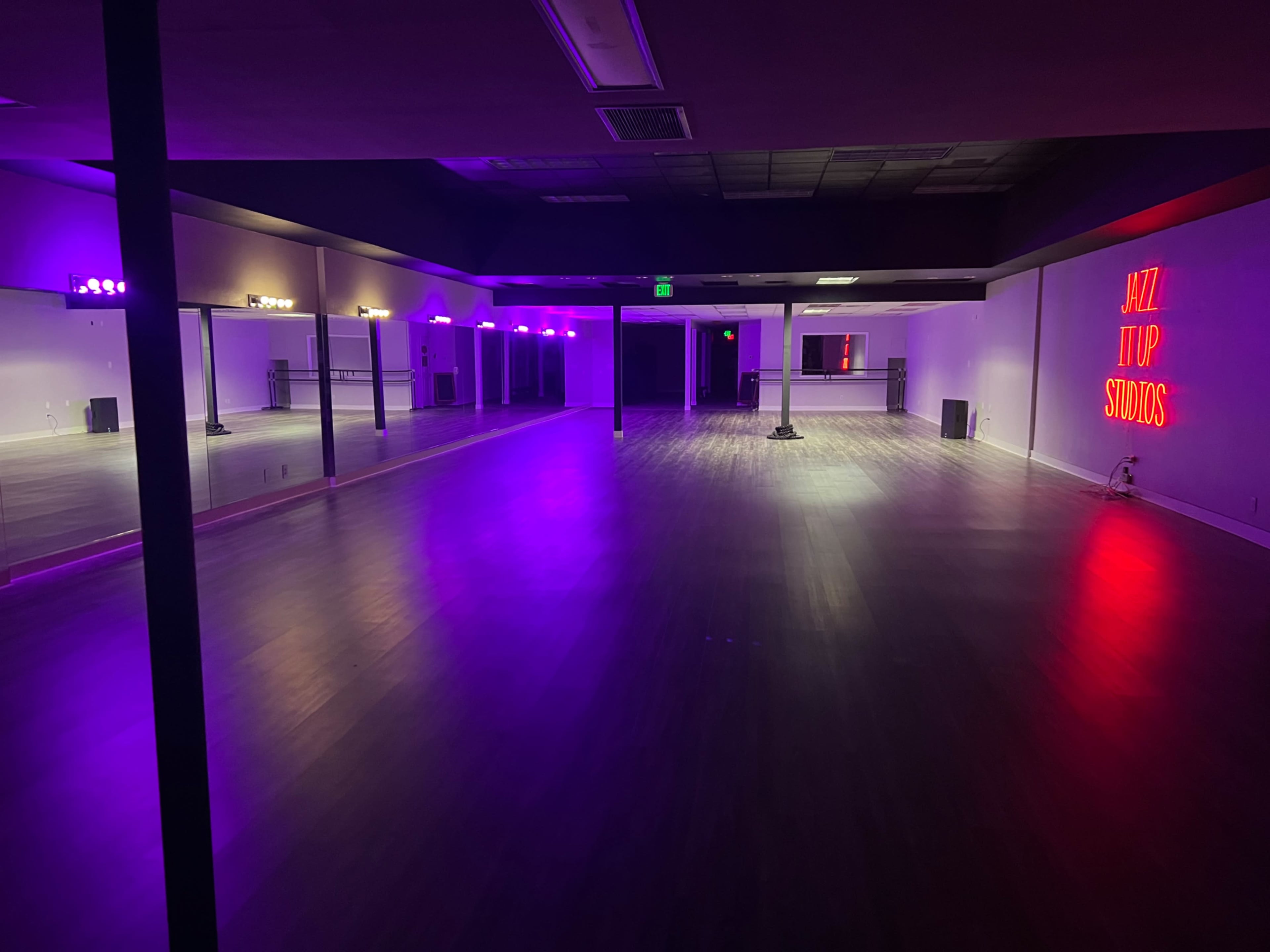 A spacious dance studio with a wooden floor, illuminated by colorful lights and featuring a neon sign that reads "JAZZ IT UP STUDIOS."