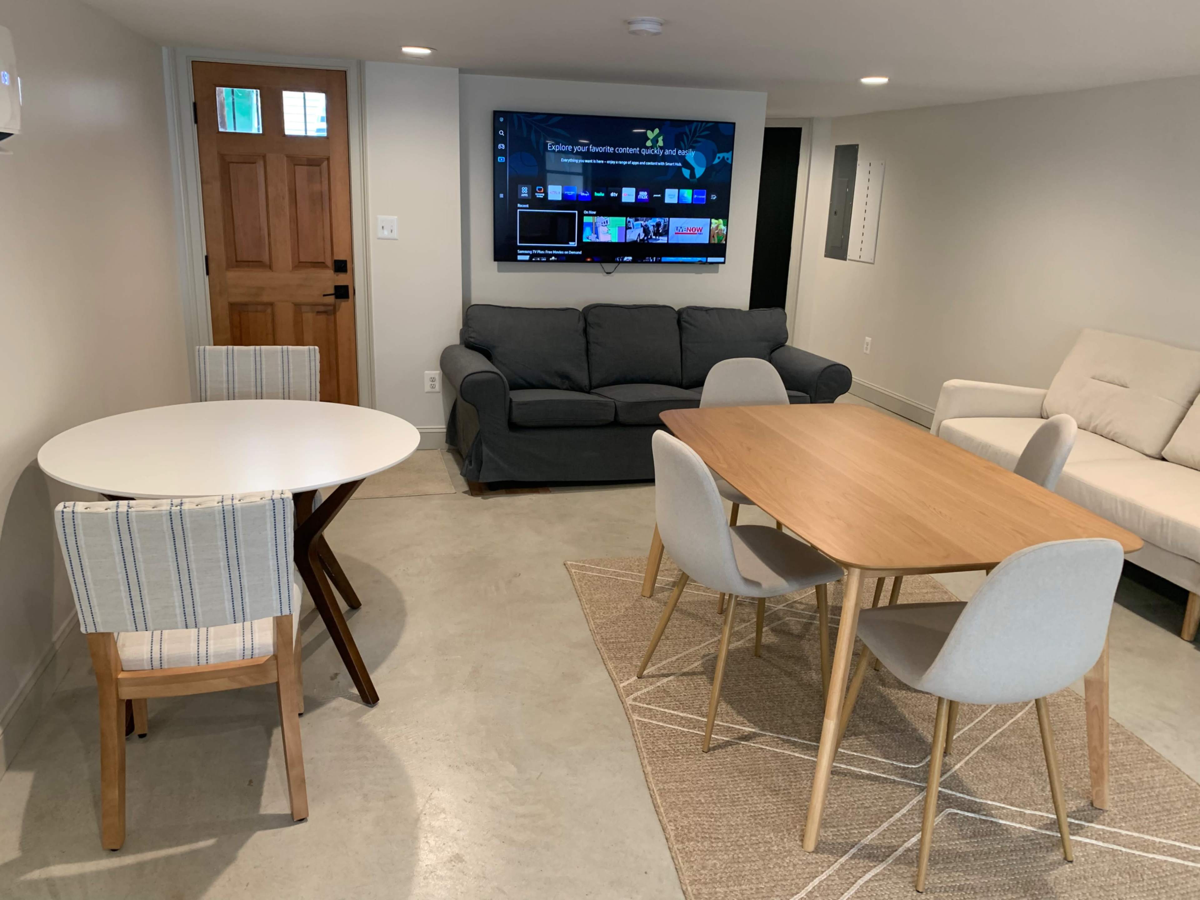 A modern basement living area featuring two seating areas, a television, a round table with striped chairs, and a rectangular table with light-colored chairs.