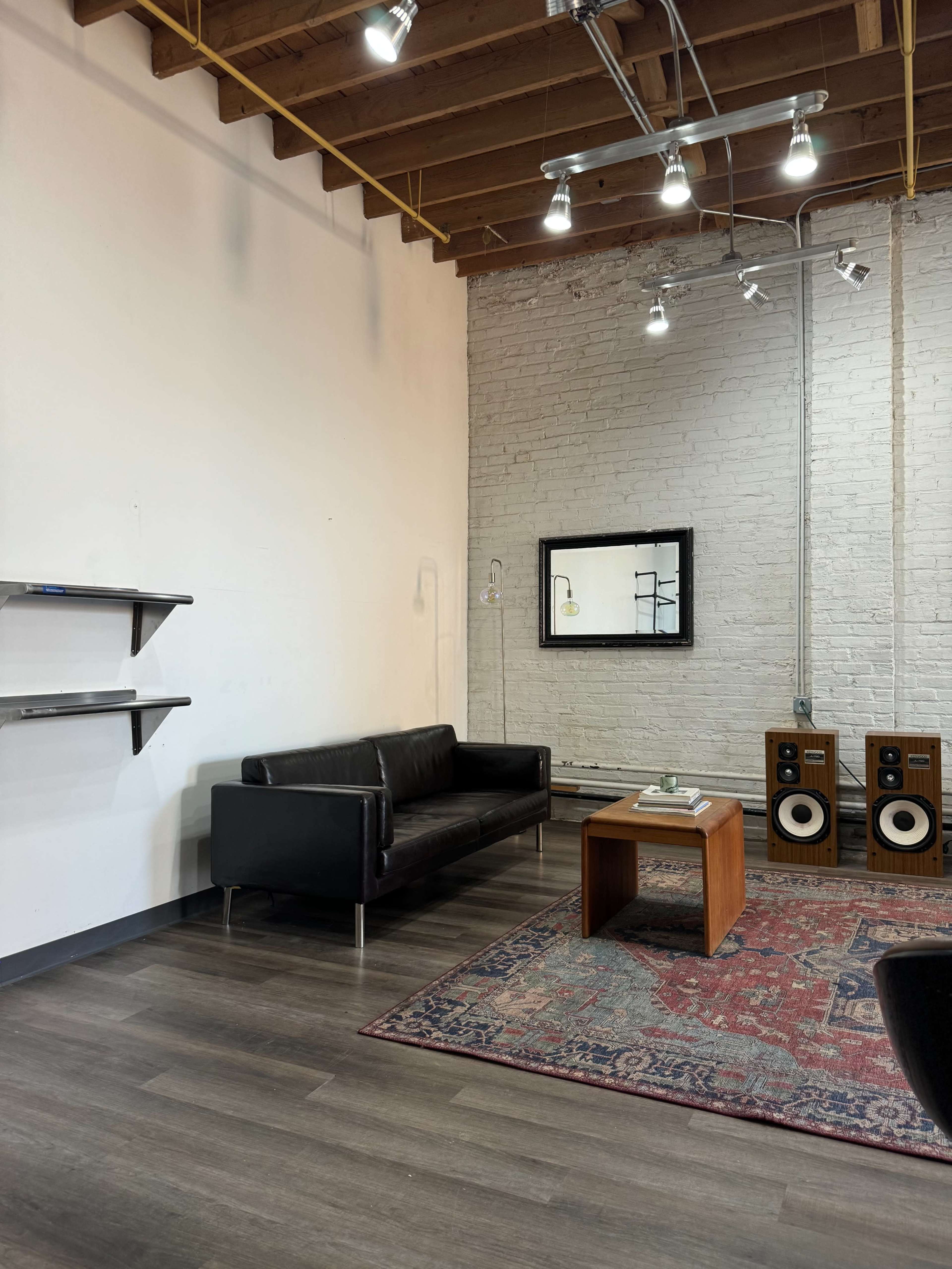 The image shows a minimalist living room with a black sofa, a wooden coffee table, a decorative rug, and audio speakers against a white brick wall.
