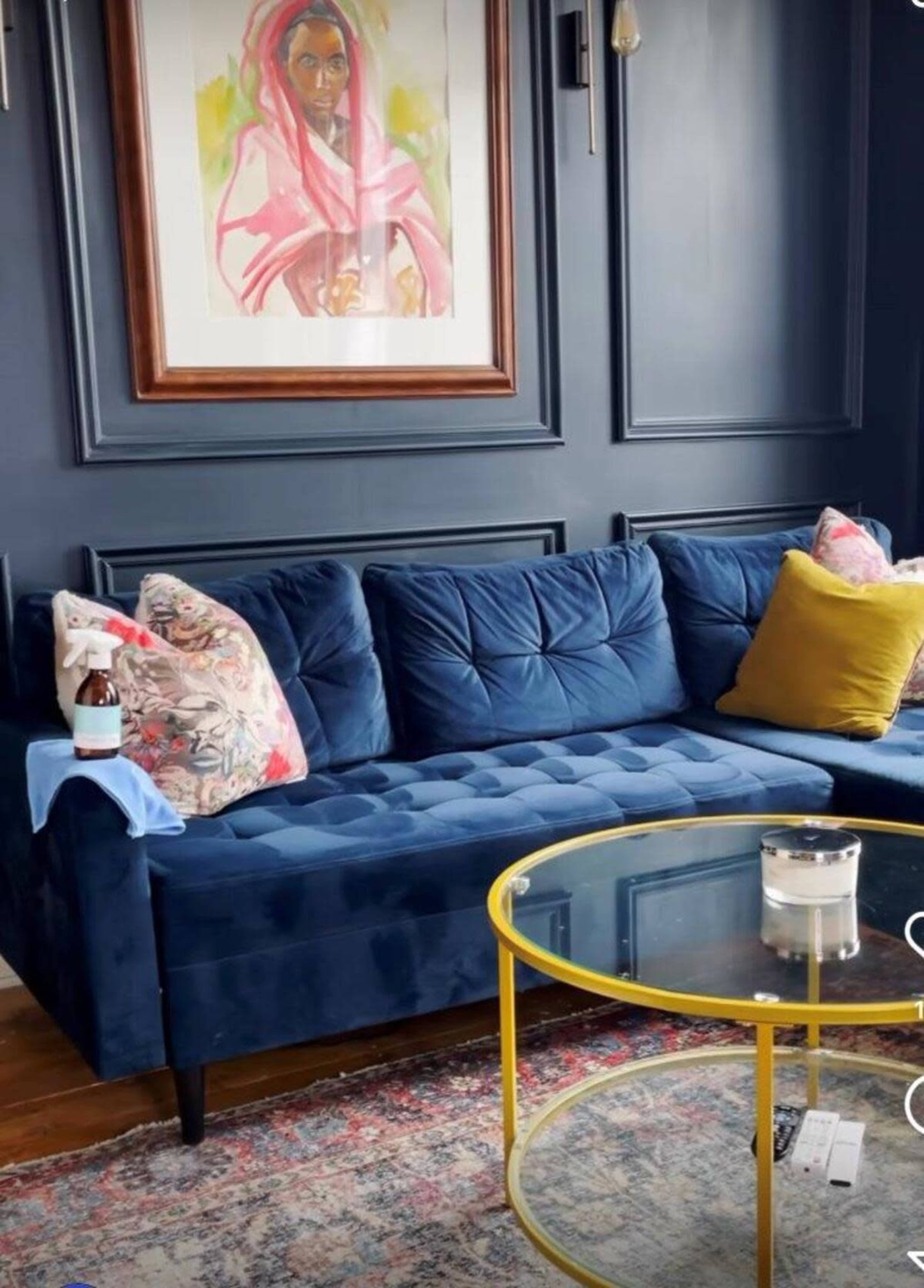 A blue velvet sectional sofa with decorative pillows, a round glass coffee table, and a colorful artwork on the wall.