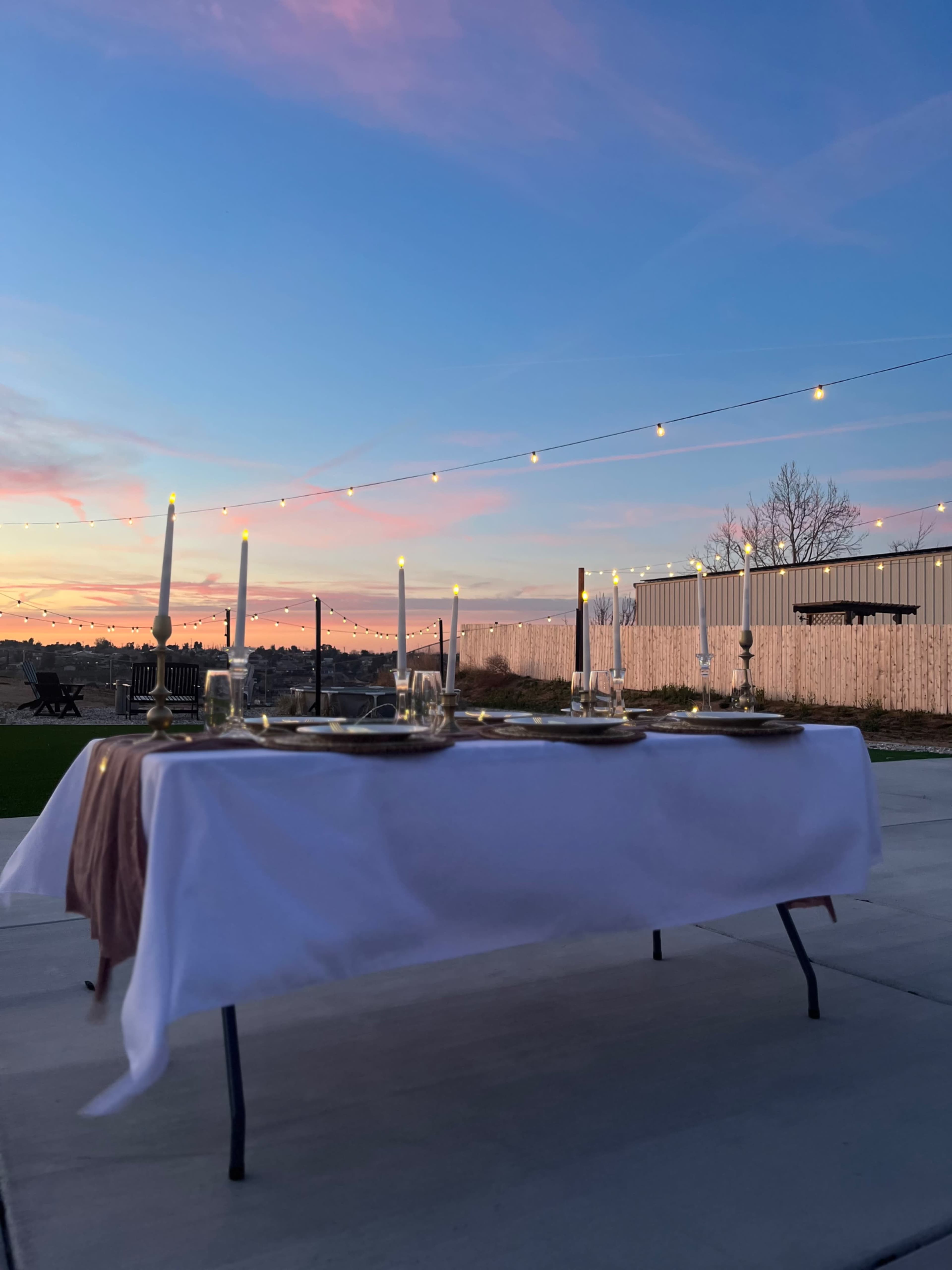A table set with candles and dinnerware is positioned outdoors under a colorful sunset and string lights.