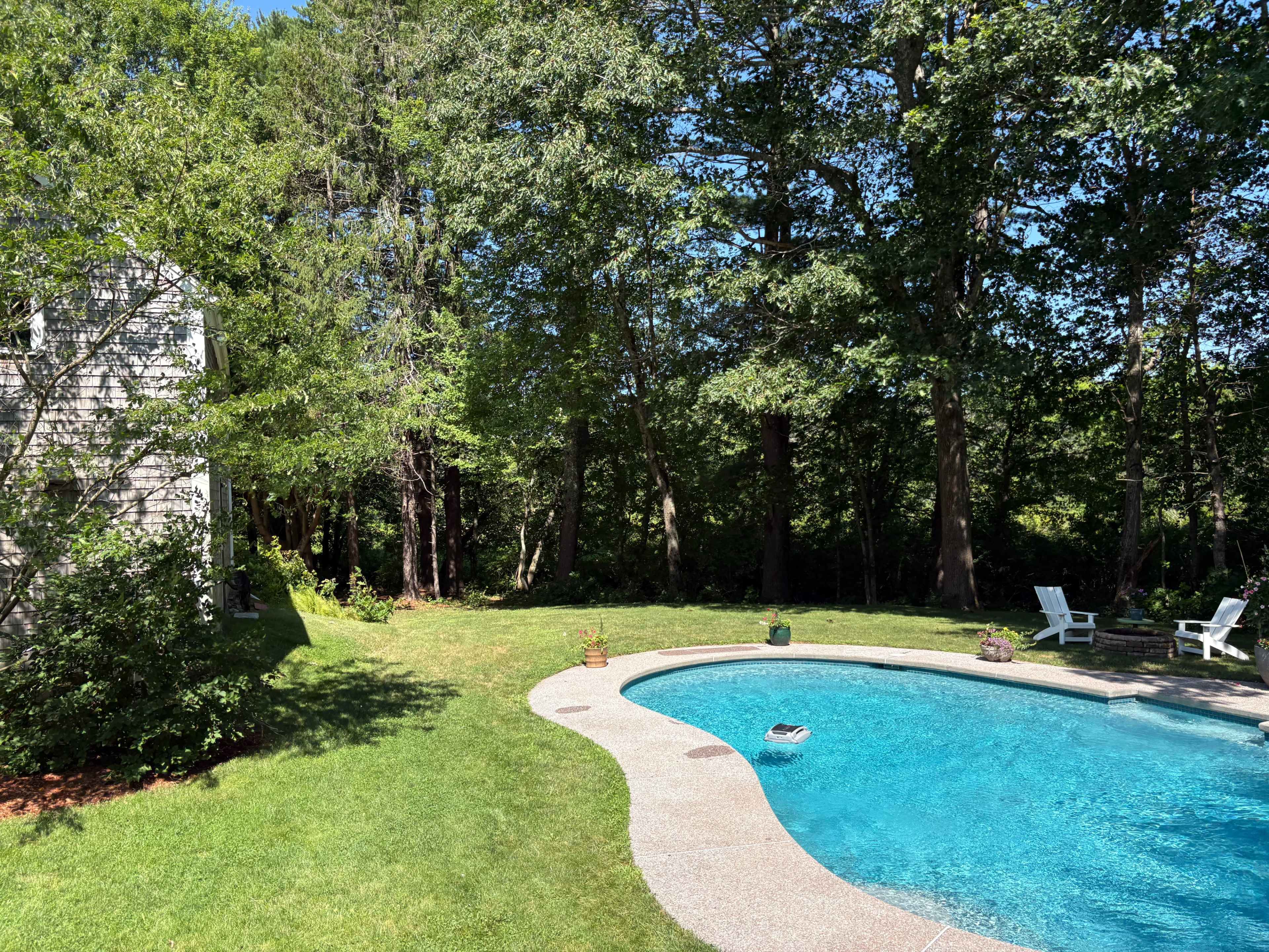 The image shows a clear swimming pool surrounded by a grassy area and trees, with a house partially visible on the left side.