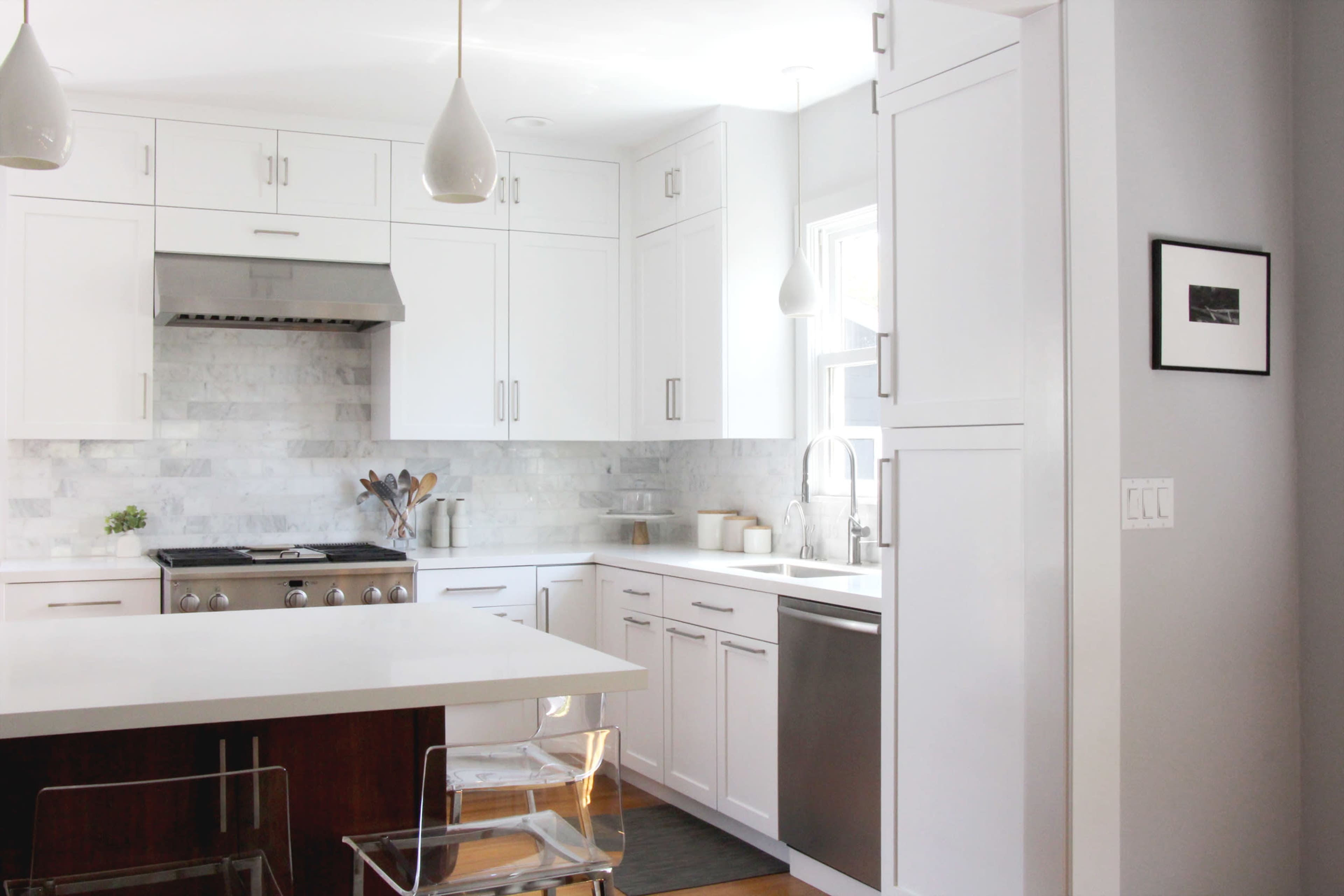 A modern kitchen features white cabinets, a stainless steel stove, and a marble backsplash, with a central island and transparent chairs.