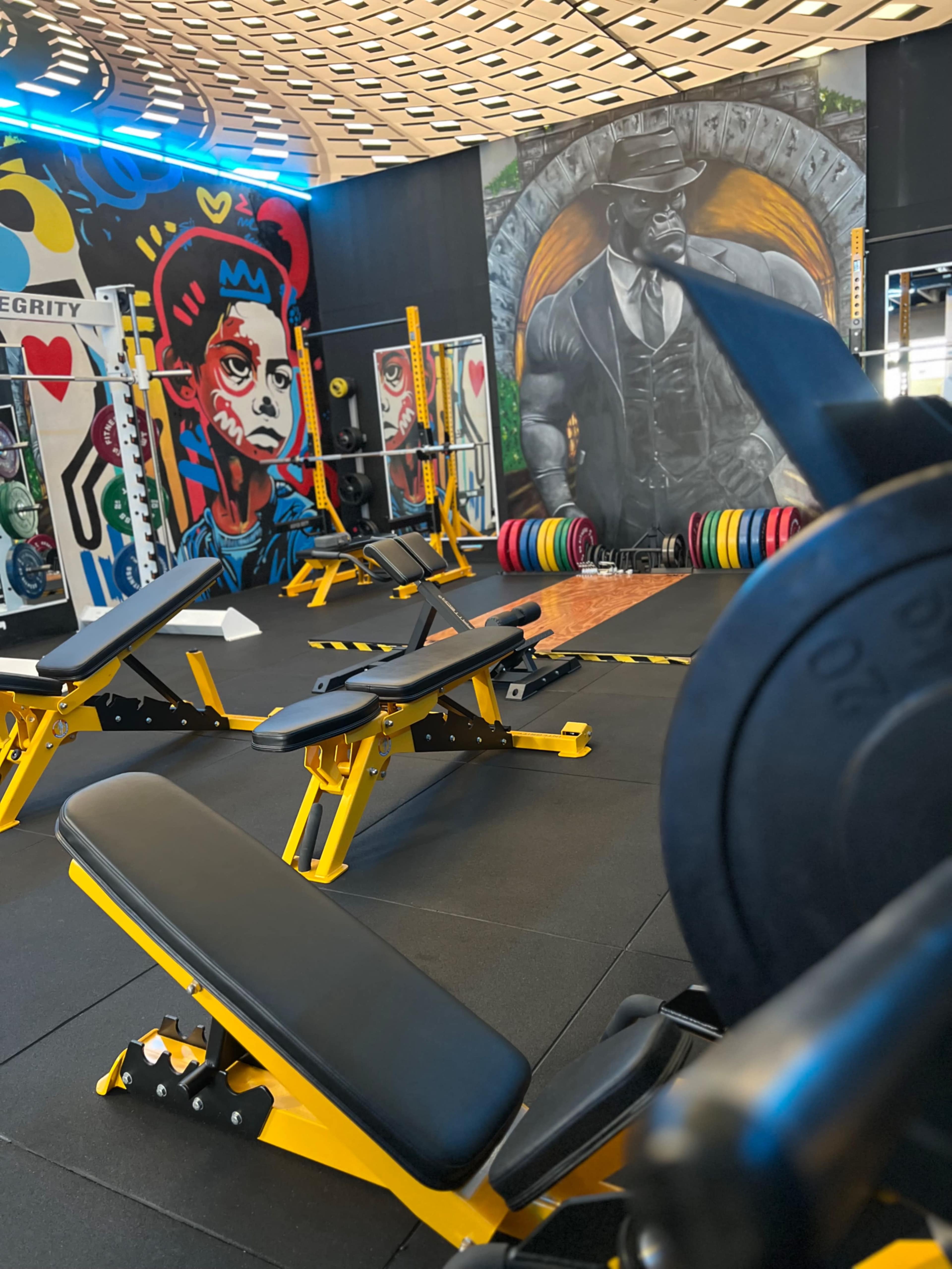 The gym features weight benches and a colorful mural of a muscular figure alongside various weight plates.